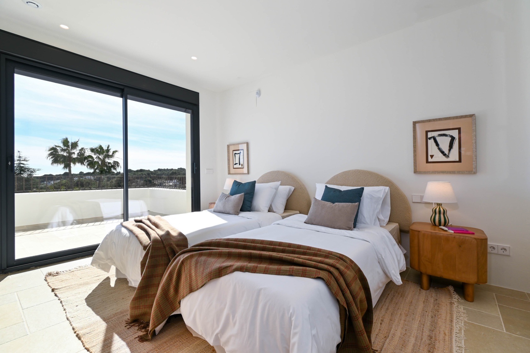Dormitorio luminoso con dos camas individuales, cabeceros curvos y acceso directo a una terraza privada con vistas a las palmeras y el entorno natural.