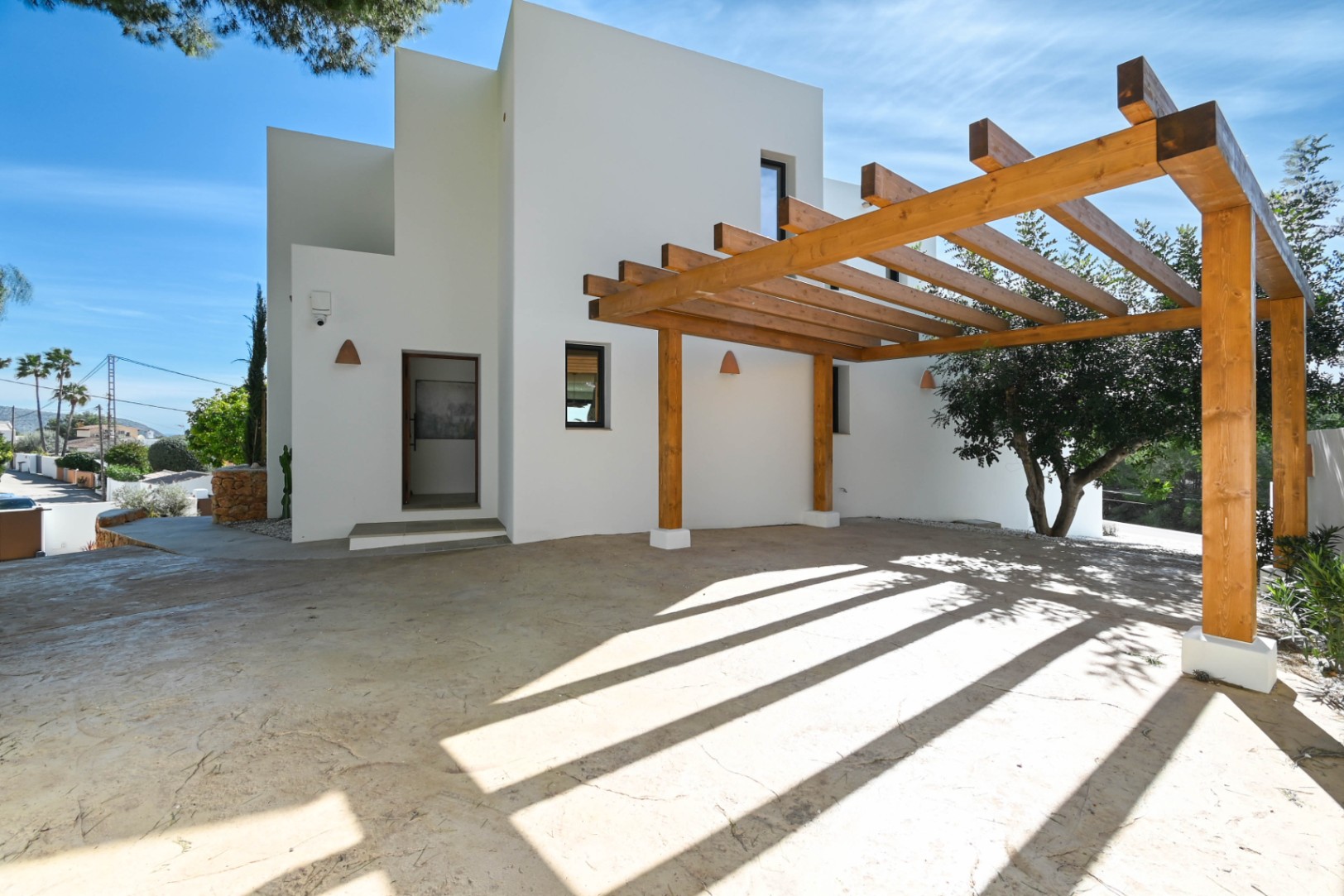 Fachada minimalista blanca con pérgola de vigas de madera natural sobre zona de aparcamiento pavimentada. Incluye apliques de pared cerámicos y acceso principal.