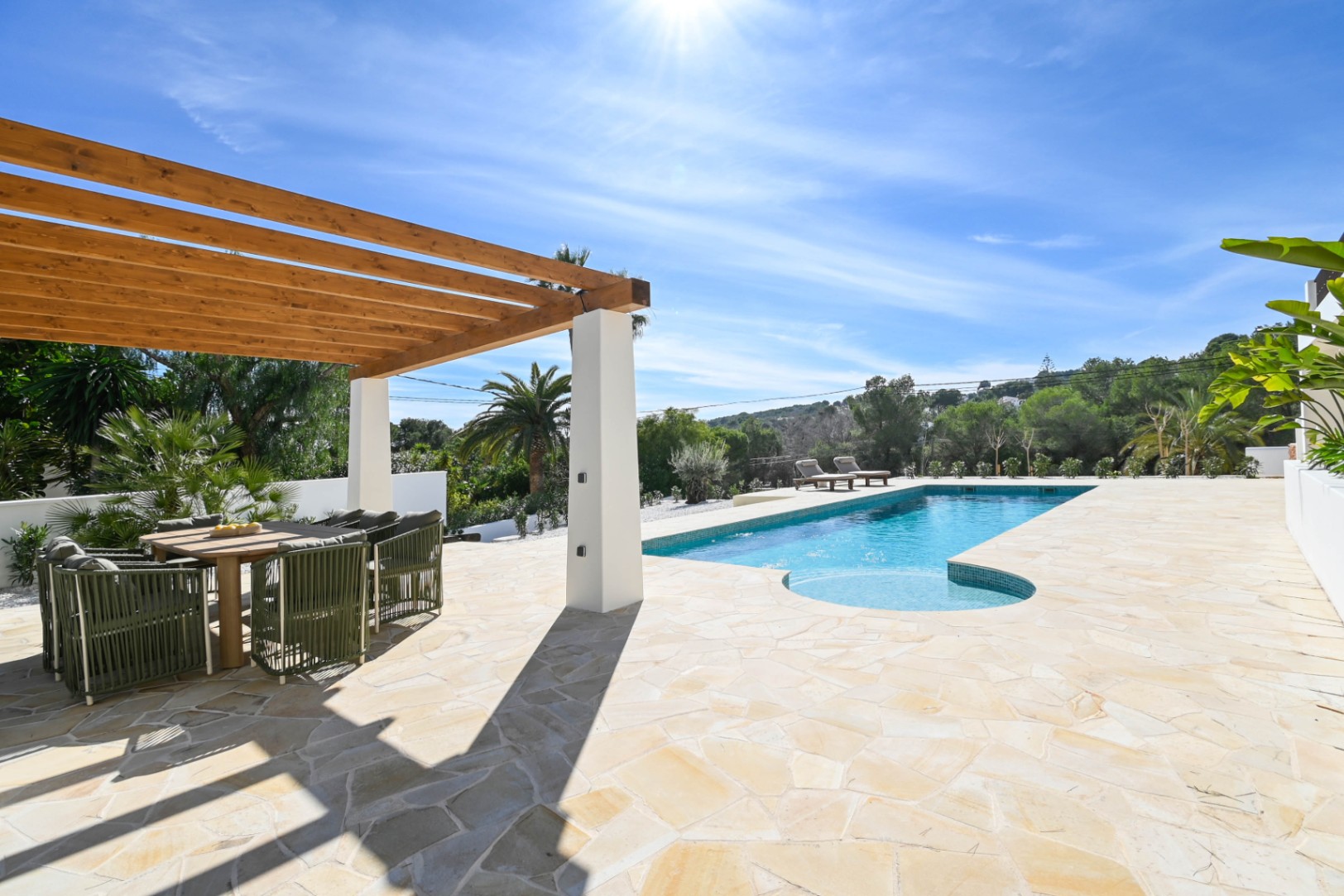 Terraza exterior con pavimentación de piedra natural, pérgola de madera con comedor y una piscina de 13 metros rodeada de vegetación mediterránea.