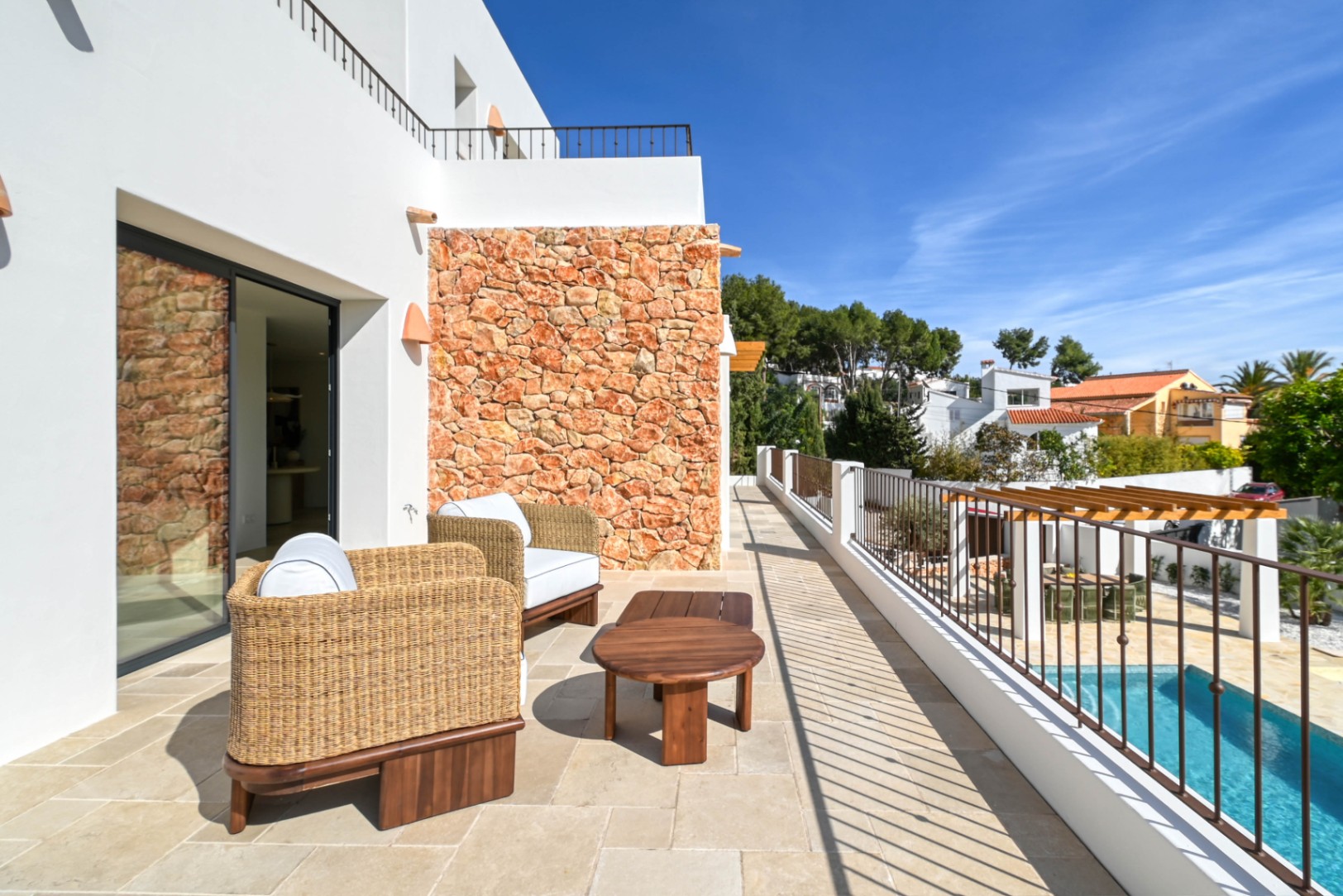 Terraza superior con suelos de piedra, mobiliario de mimbre y un muro de mampostería natural, con vistas despejadas hacia la piscina y la zona de comedor exterior.