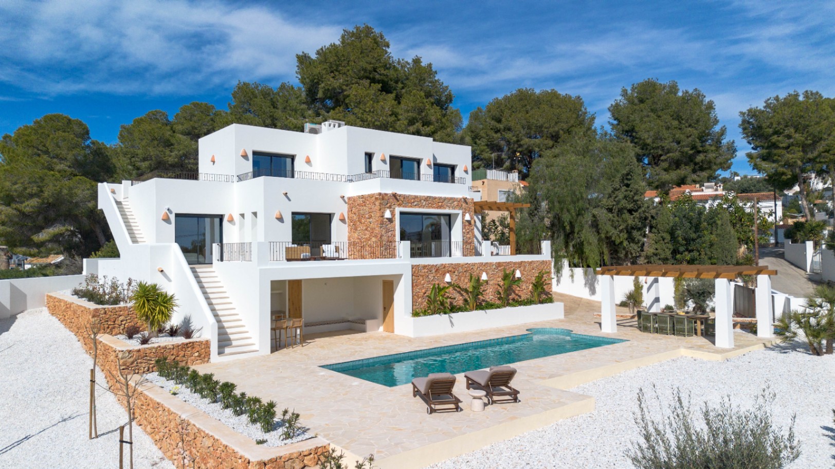 Vista exterior de la villa con fachada blanca y muros de piedra natural, destacando la piscina, terrazas en varios niveles y zona de comedor bajo pérgola.