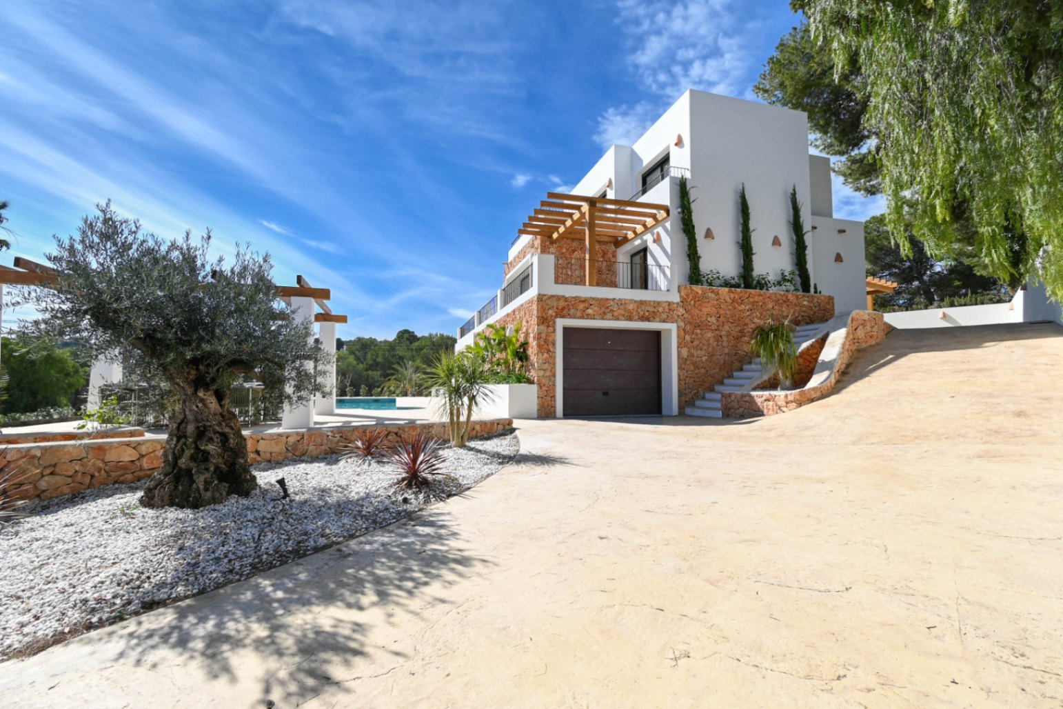 Vista exterior de la villa mostrando la fachada de piedra natural, el garaje integrado y el acceso pavimentado rodeado de olivos y vegetación mediterránea.