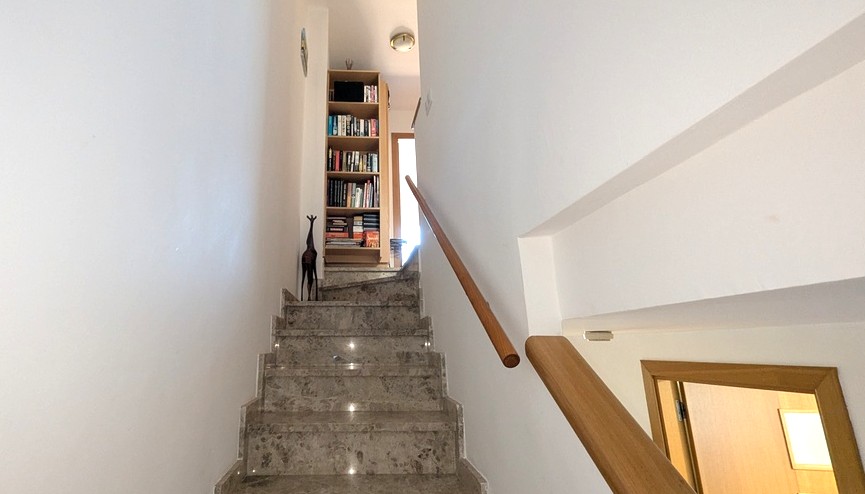 Escalera interior de mármol con pasamanos de madera que comunica las plantas, destacando una librería integrada en el rellano superior.