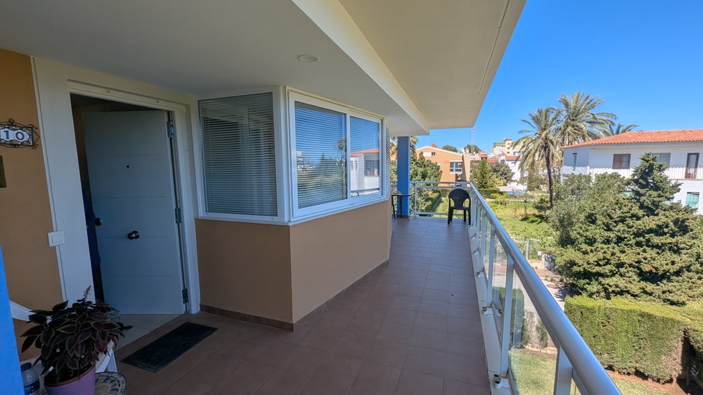 Terraza de entrada con suelo de gres y barandilla de cristal, con vistas a la vegetación comunitaria y villas mediterráneas de Jávea bajo un cielo despejado.