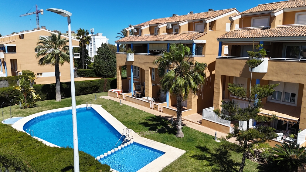 Vista elevada de la zona de piscina comunitaria y césped, mostrando la fachada en tonos terracota con pilares azules y terrazas privadas del complejo.