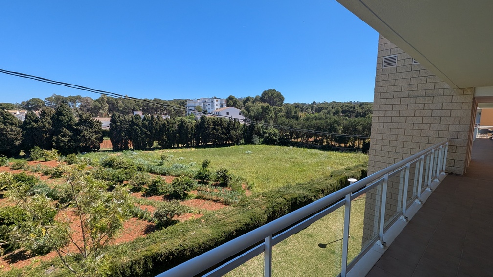 Vistas desde la terraza con suelo cerámico y barandilla de cristal, hacia paisajes verdes despejados y una hilera de cipreses bajo el cielo azul.