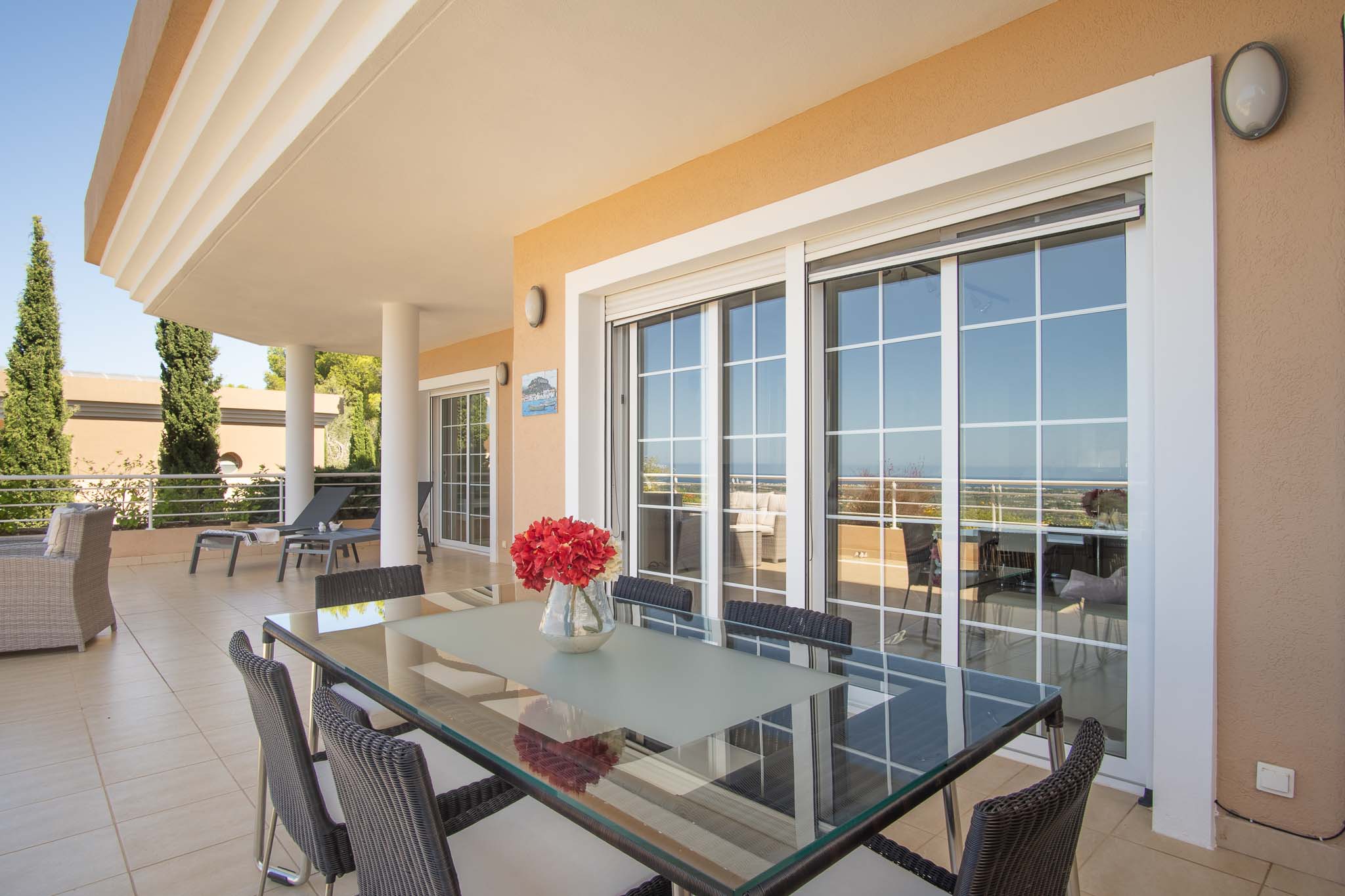 Amplia terraza cubierta con comedor exterior, mesa de cristal y sillas de mimbre, conectada al interior mediante grandes ventanales con vistas al mar.