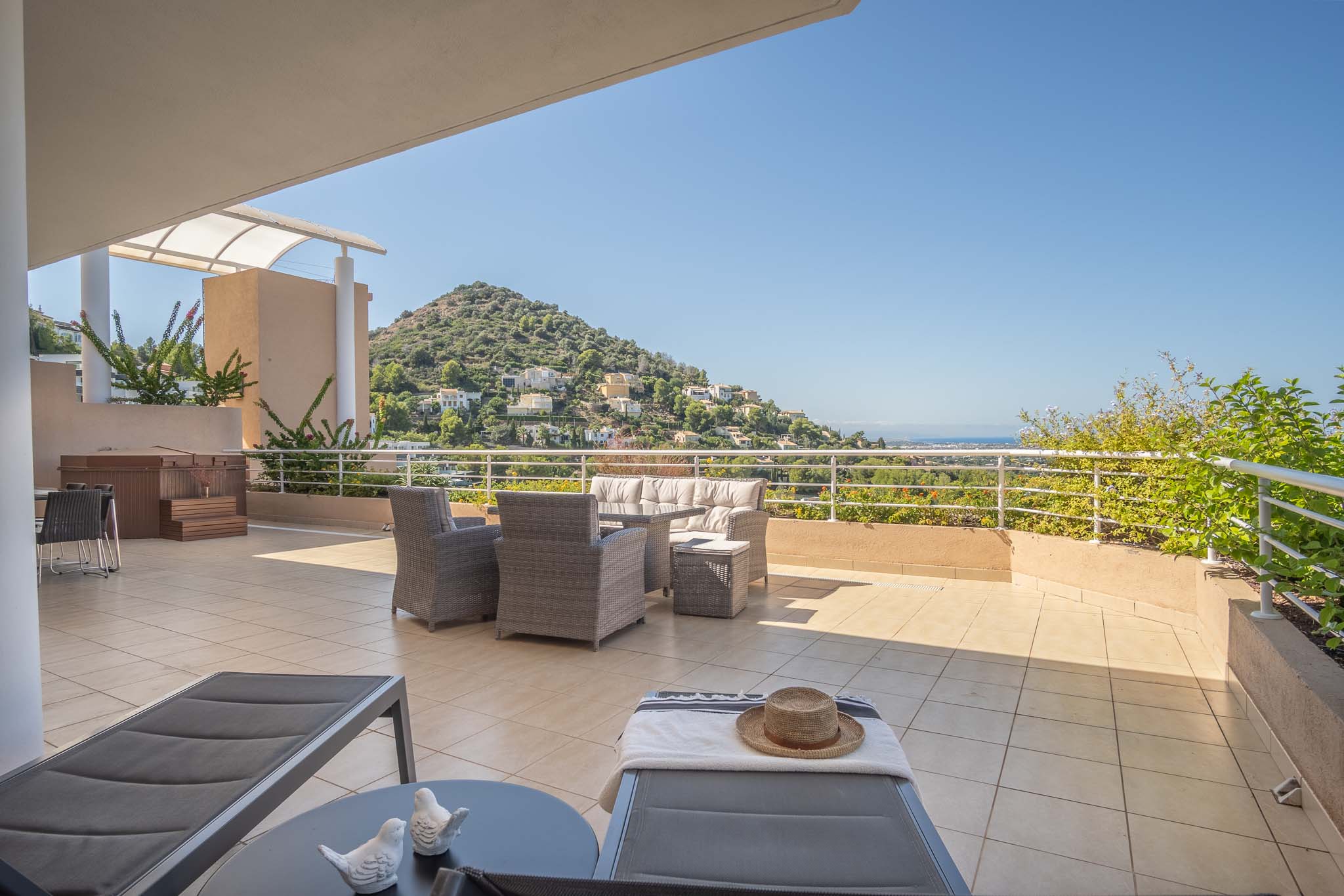 Amplia terraza perimetral con suelo cerámico, mobiliario de mimbre gris y jacuzzi privado. Ofrece vistas despejadas a la montaña y al mar Mediterráneo bajo luz natural.