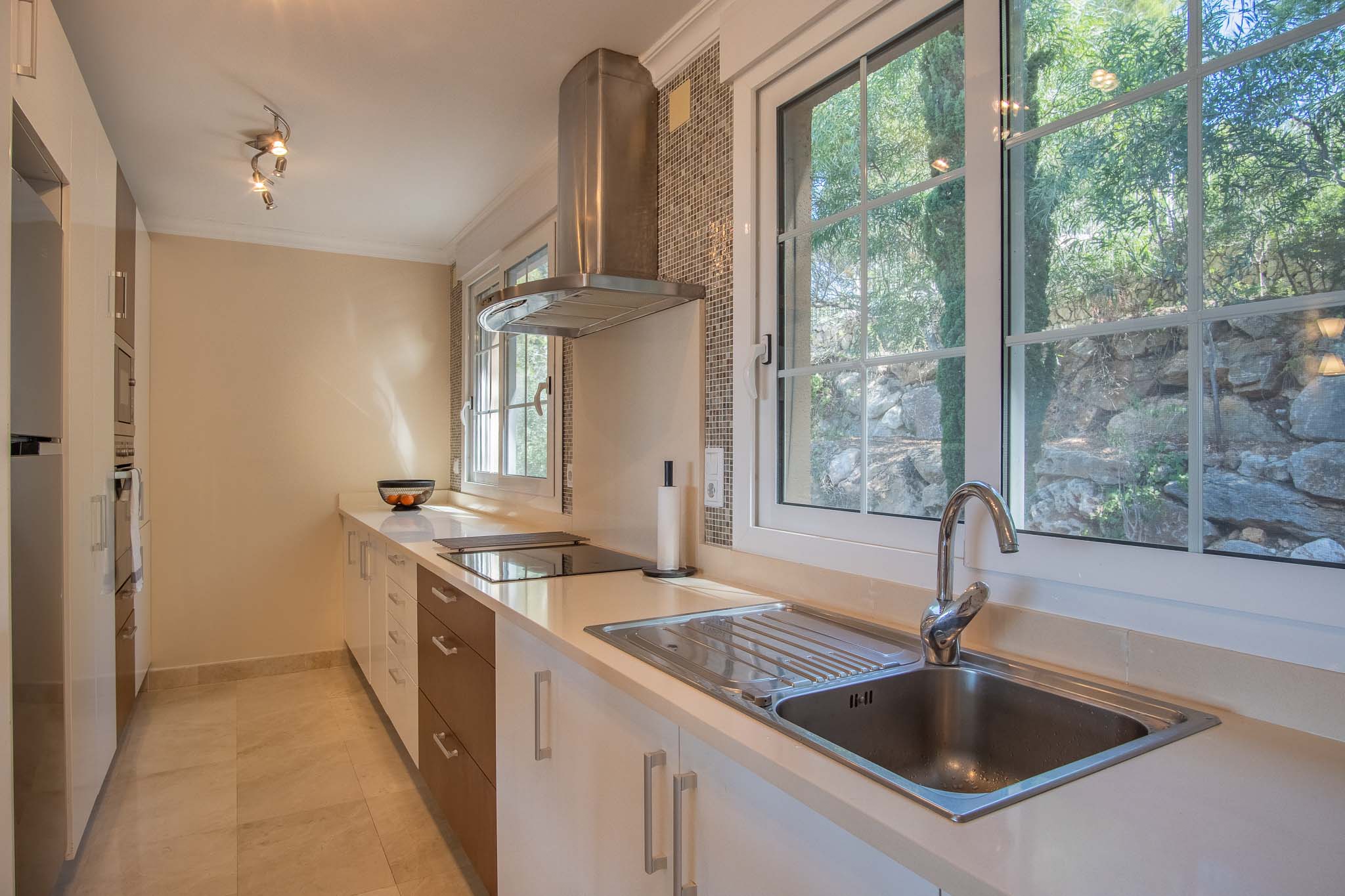 Cocina funcional con encimera blanca, campana de acero inoxidable y amplios ventanales que aportan luz natural y vistas al jardín de piedra.