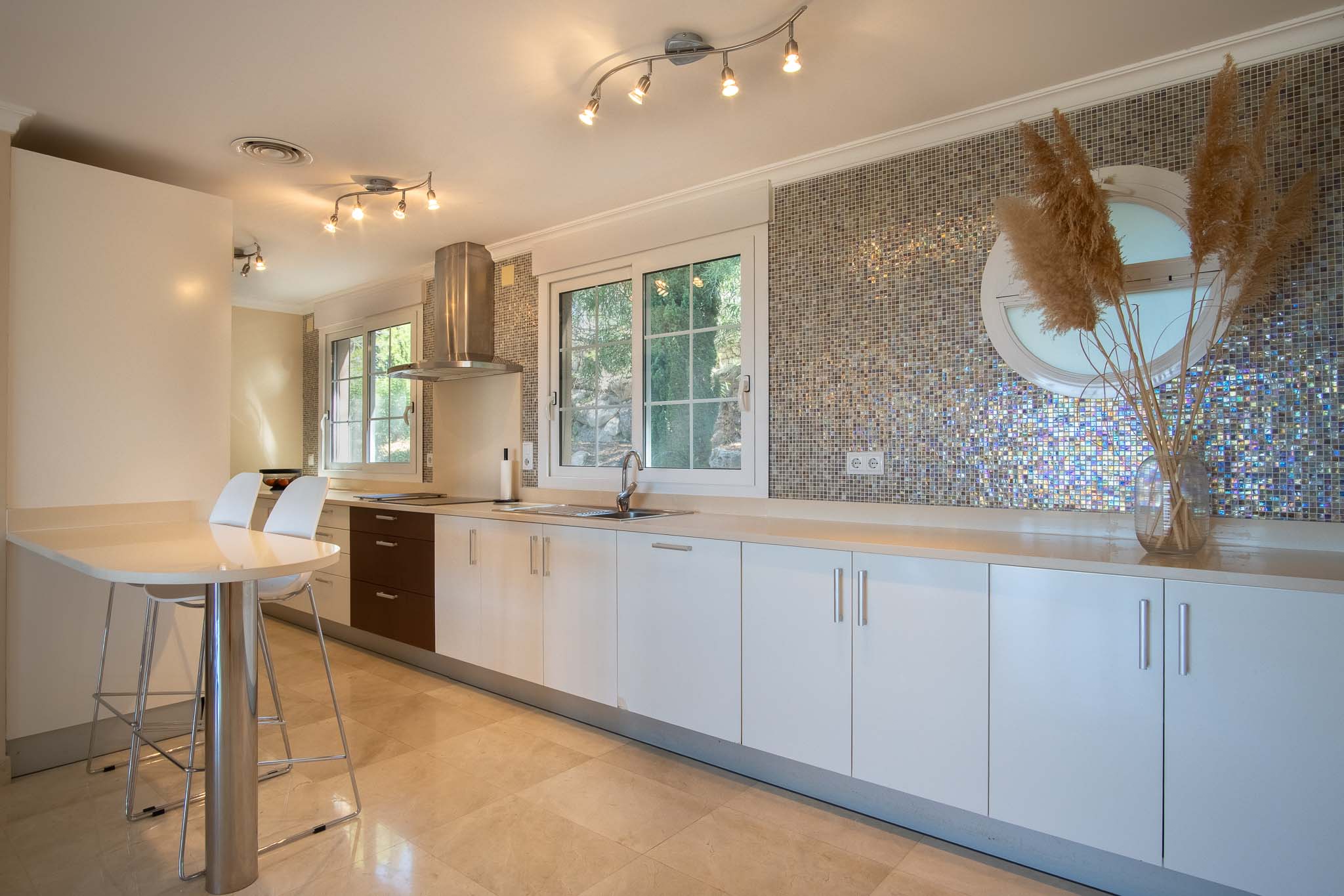 Cocina moderna con armarios blancos, encimera clara y un distintivo frente de mosaico iridiscente. Incluye mesa de desayuno, suelos de mármol y ventanas que aportan luz natural.