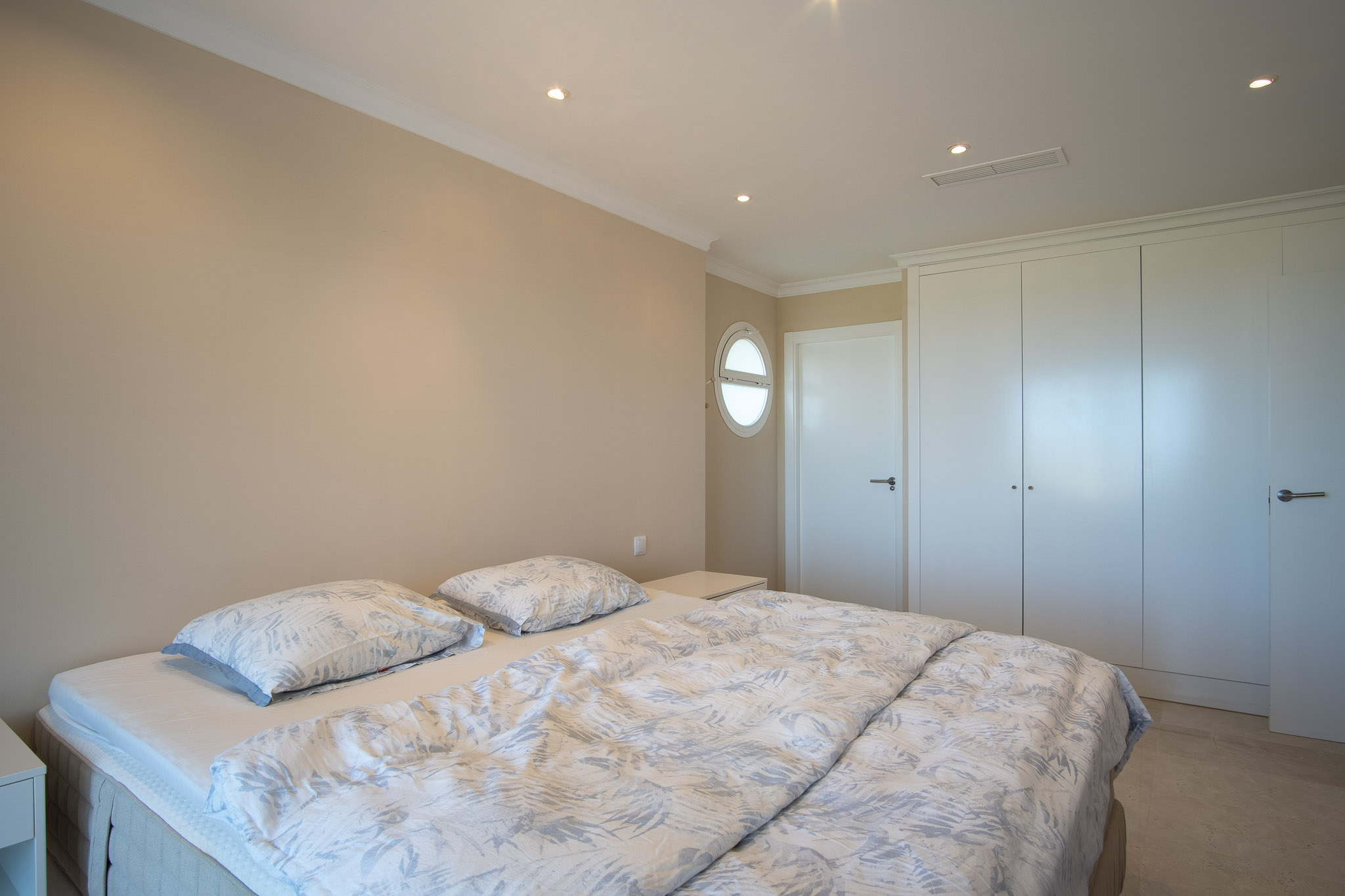 Dormitorio con cama doble, armarios empotrados blancos y suelos de mármol. Incluye iluminación empotrada y una ventana circular tipo ojo de buey.