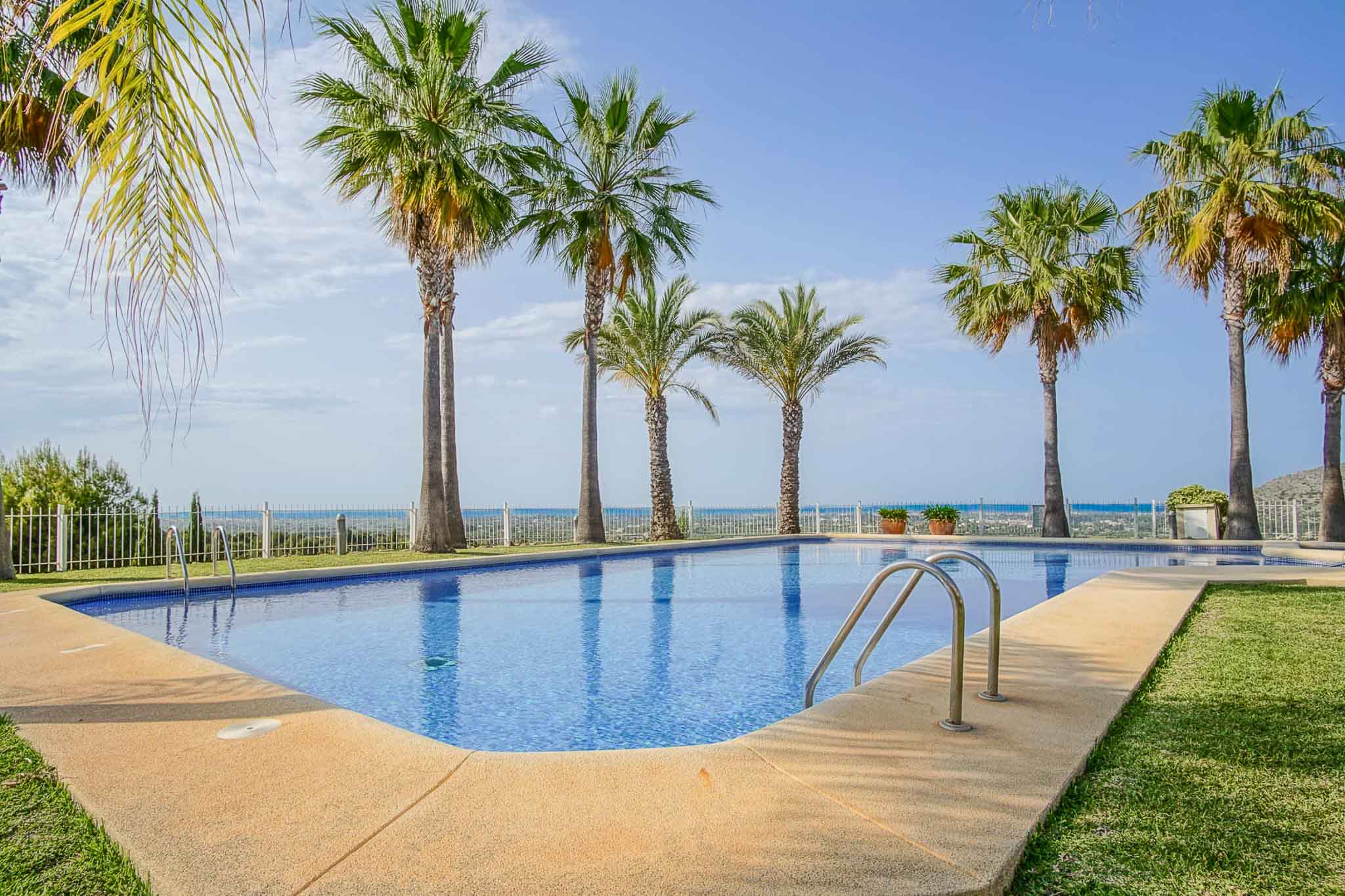 Piscina comunitaria rectangular con revestimiento de gresite azul, rodeada de una playa de piedra clara, césped y palmeras con vistas panorámicas al mar Mediterráneo.