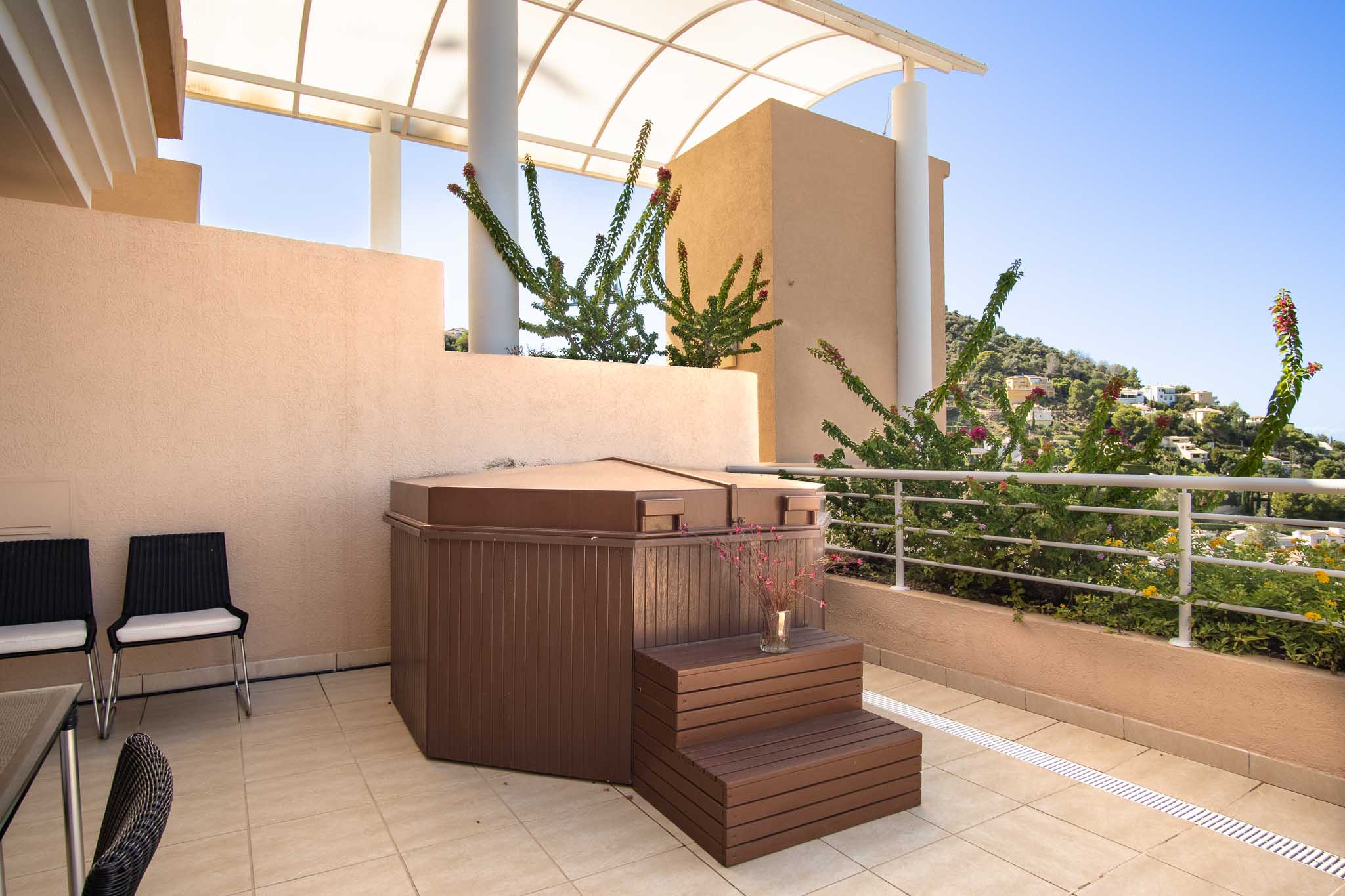 Terraza privada con jacuzzi cubierto y escalones de madera, bajo una pérgola arqueada con vistas a las colinas de La Sella y vegetación mediterránea.