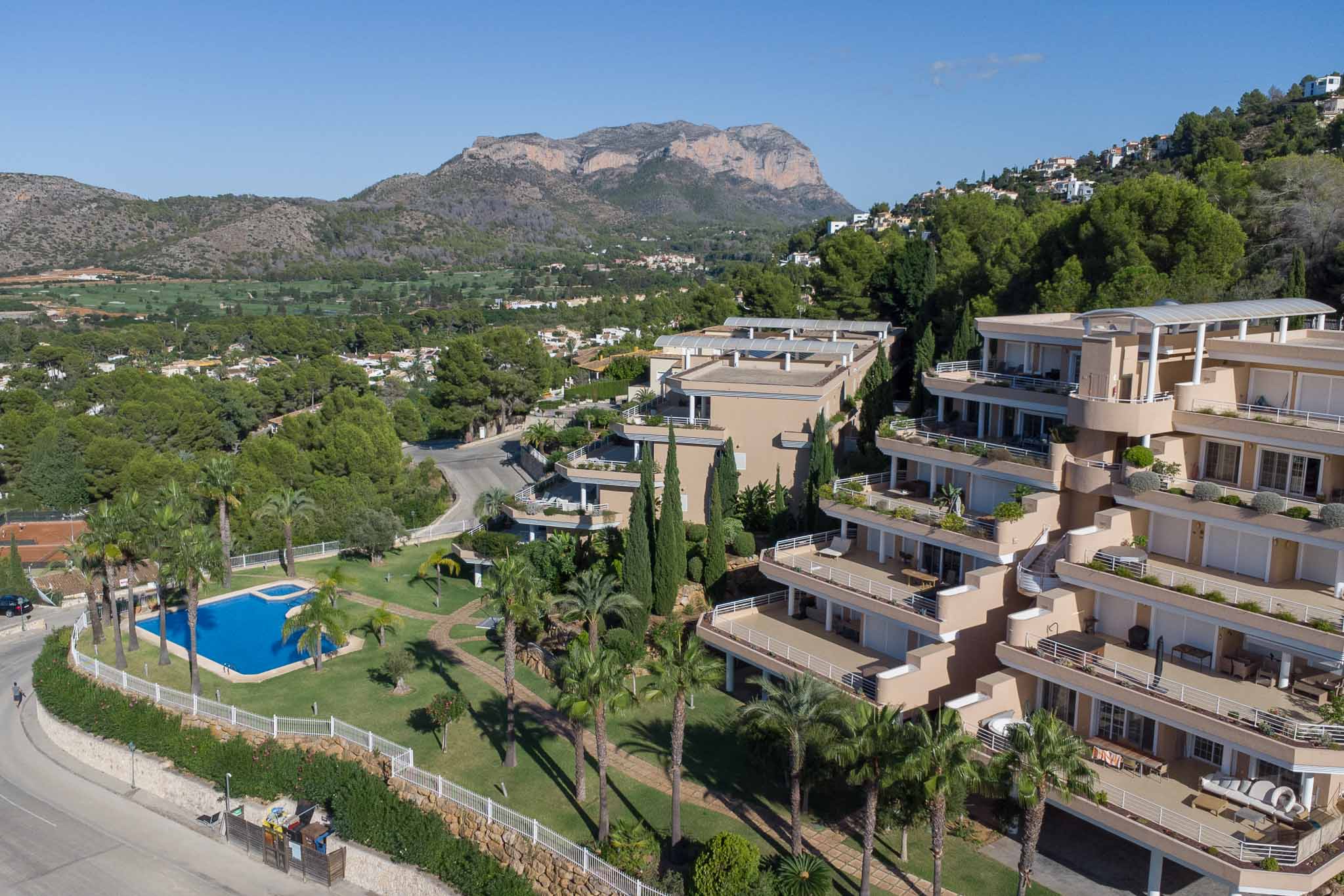 Vista aérea del complejo Las Mimosas que muestra la piscina comunitaria, jardines con palmeras y la arquitectura aterrazada frente al campo de golf y el Montgó.