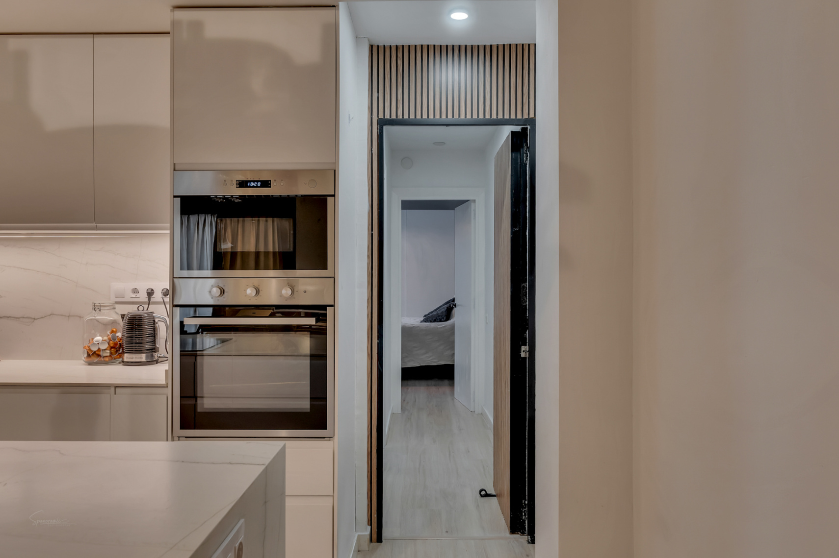 Detalle de la cocina con horno empotrado y vista al dormitorio a través de una puerta con diseño de madera.