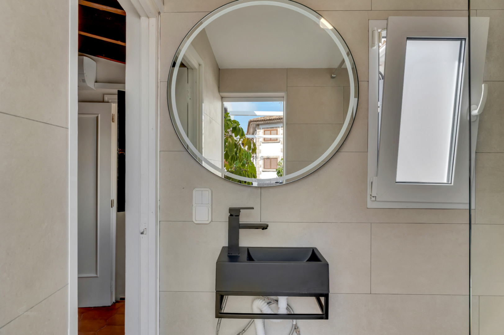 Detalle del baño con lavabo negro suspendido, espejo redondo con luz y ventana abatible.