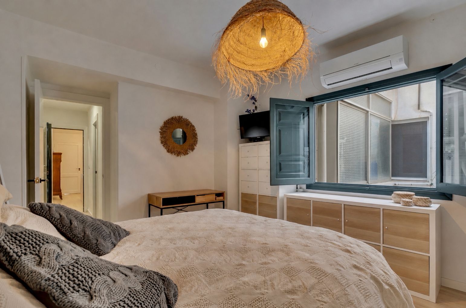 Dormitorio luminoso con cama doble, ventana de madera azul y lámpara de mimbre colgante.