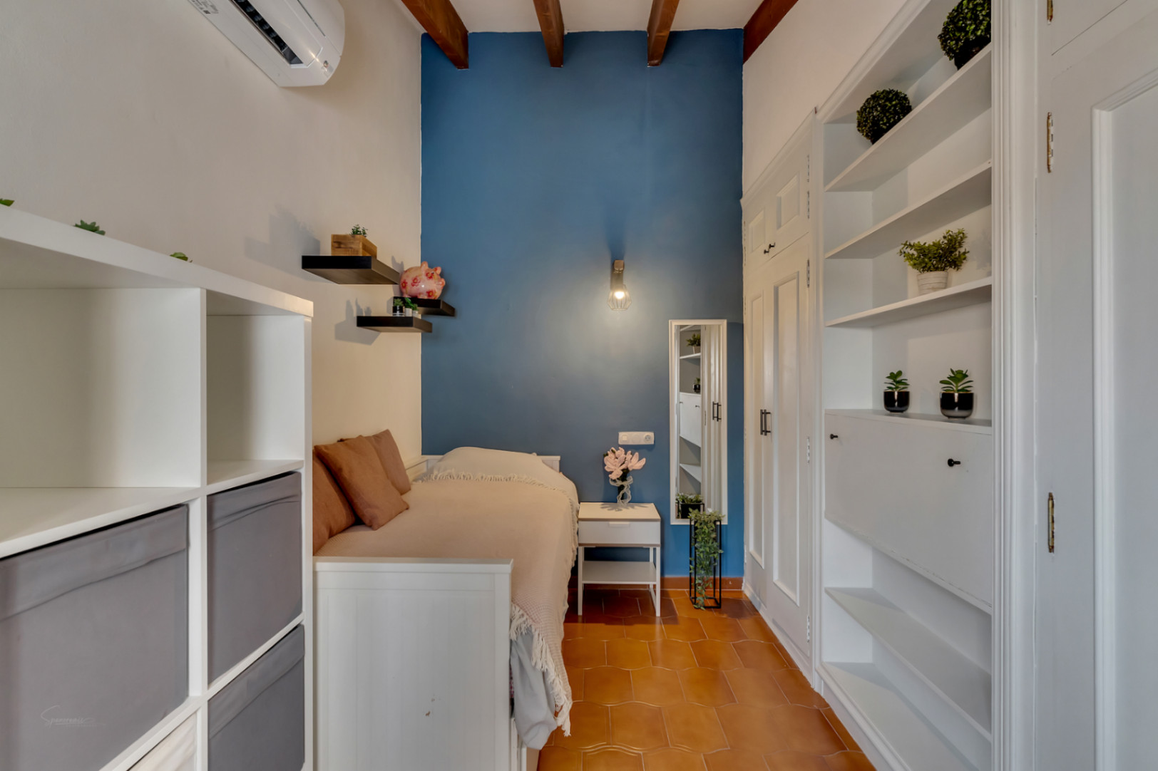Habitación con cama individual, estanterías blancas, pared azul y suelo de terracota.
