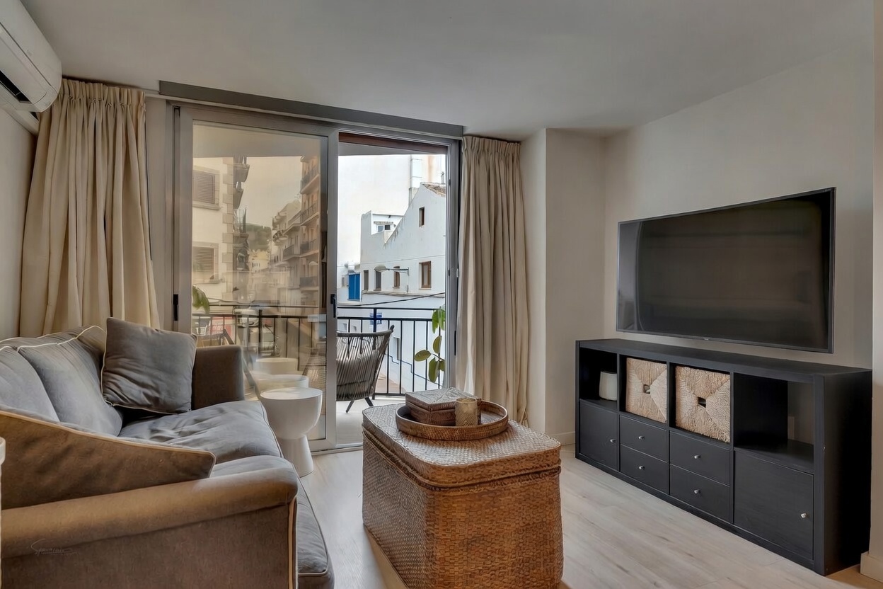Salón moderno con sofá gris, mueble de TV negro y acceso a balcón con vistas urbanas.