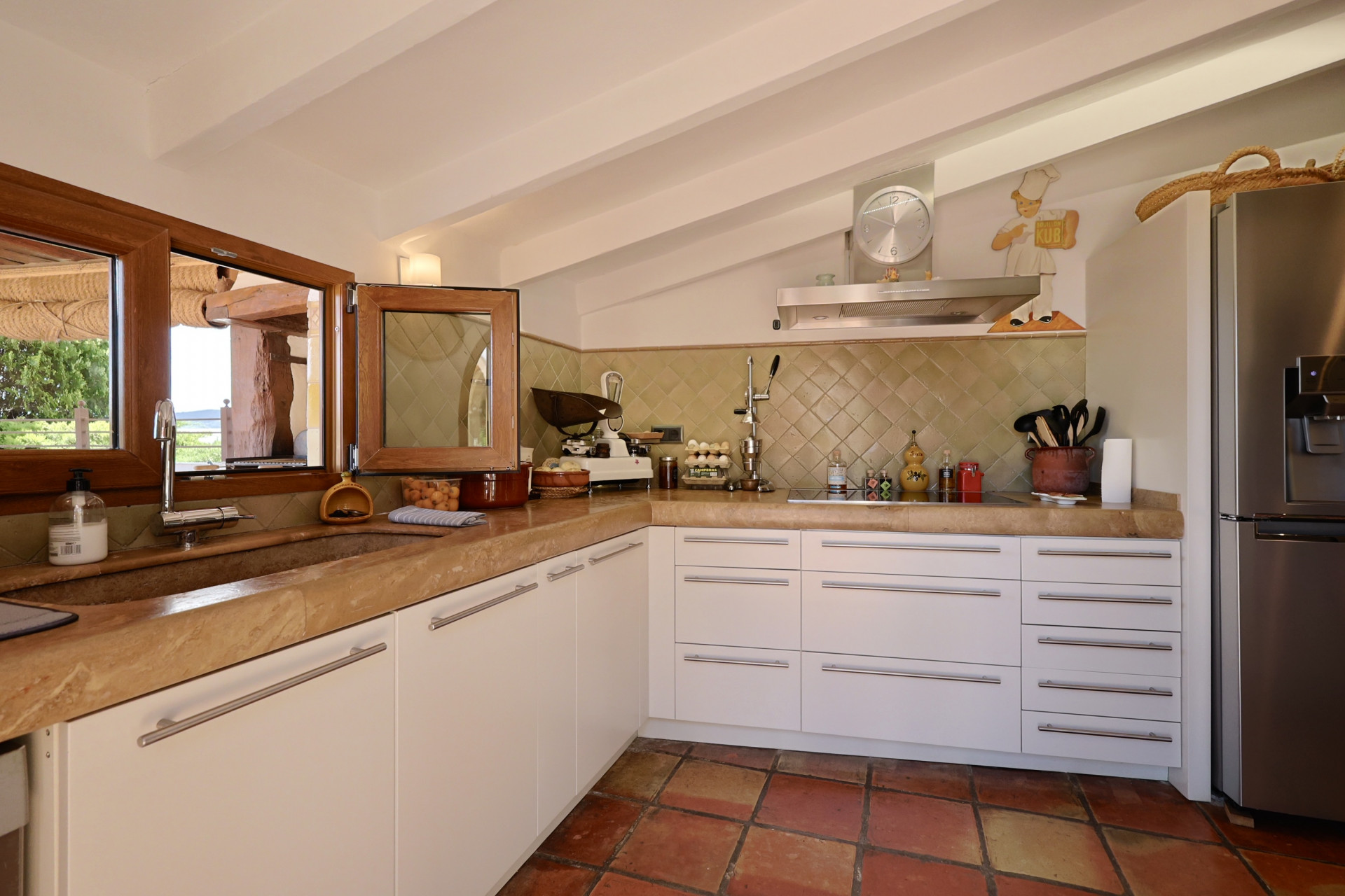 Cocina rústica con encimera de piedra, muebles blancos y suelo de terracota. Fregadero de piedra con ventana de madera.