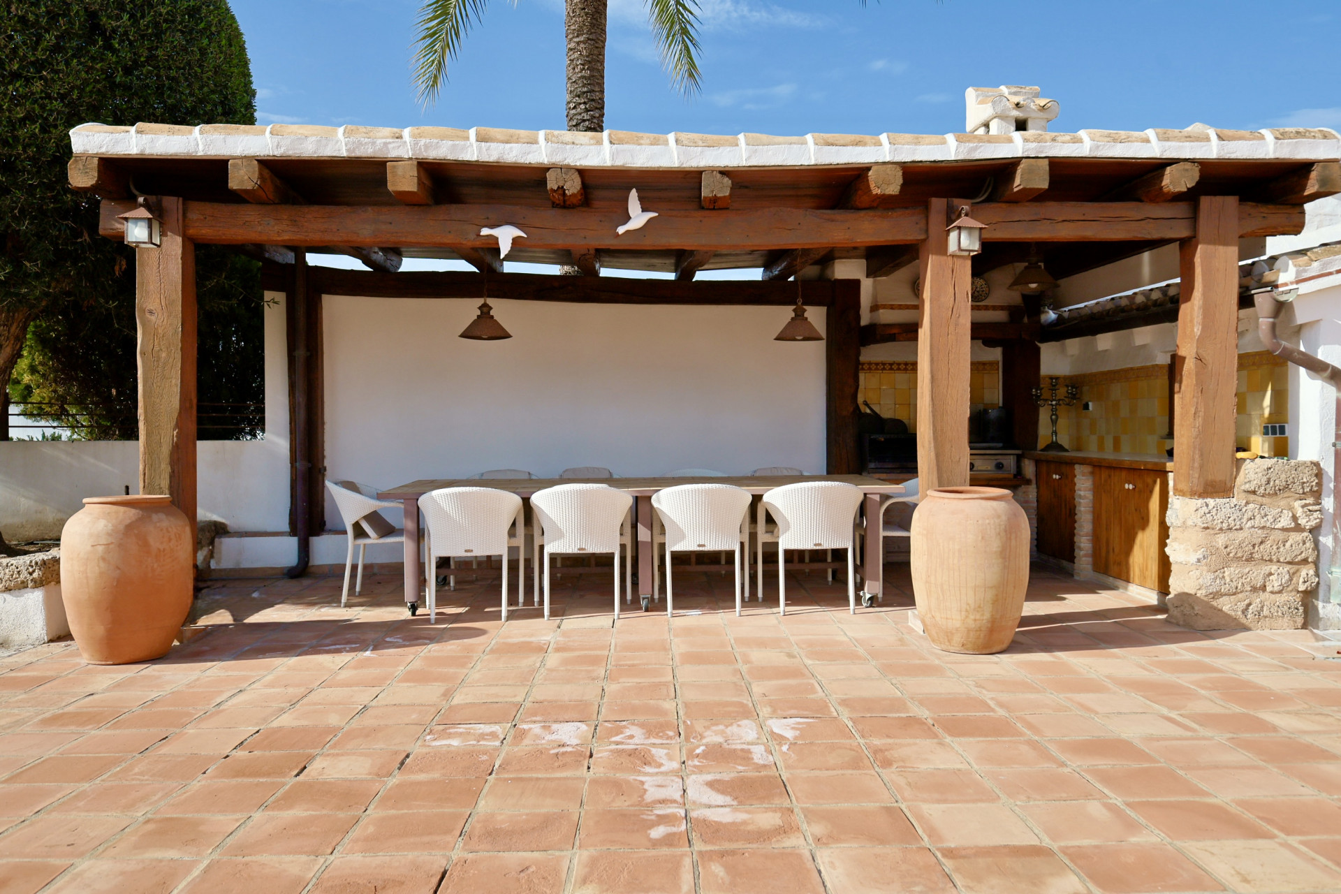 Comedor exterior cubierto con mesa larga, sillas de mimbre blancas y suelo de baldosas de terracota.