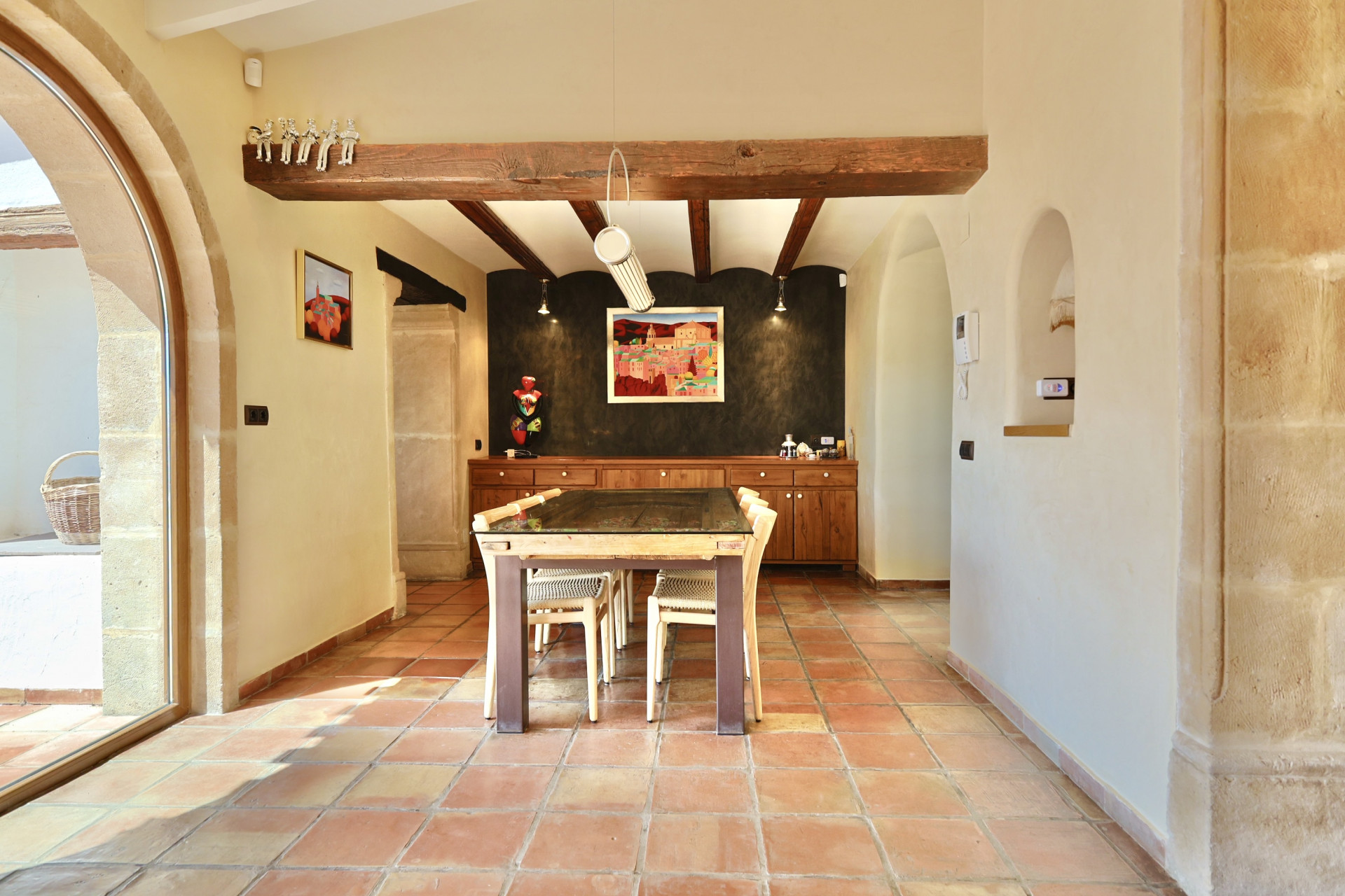 Comedor luminoso con suelo de terracota y vigas de madera, destacando una mesa con tapa de cristal.