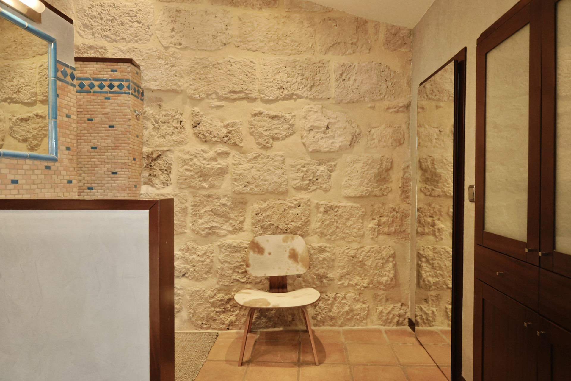 Detalle de baño con pared de piedra rústica, espejo con marco azul y silla de diseño con tapizado de piel.