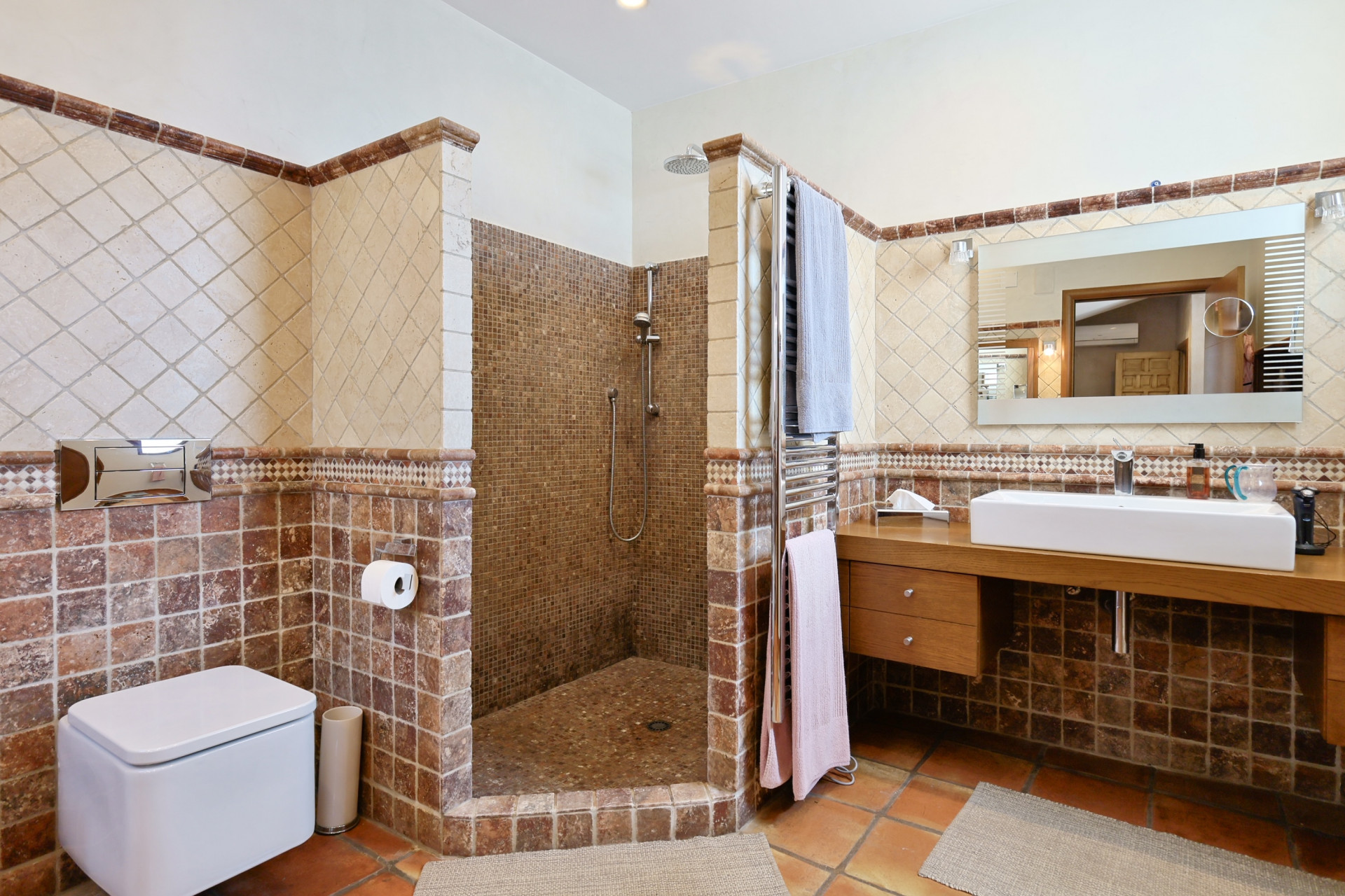 Detalle del baño con ducha de mosaico, lavabo moderno sobre encimera de madera y azulejos rústicos.