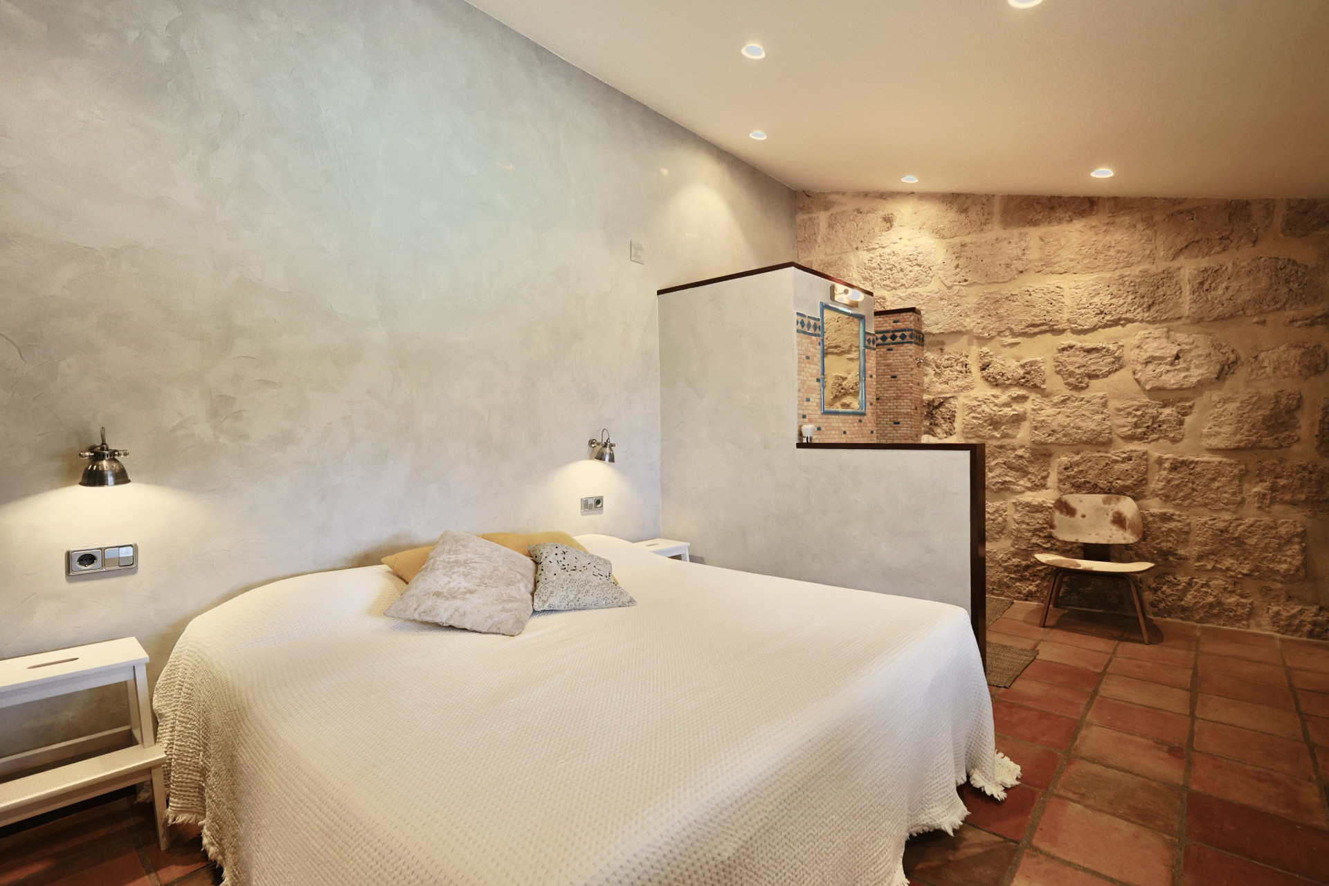 Dormitorio con pared de piedra vista, cama doble con ropa de cama blanca y suelo de terracota.