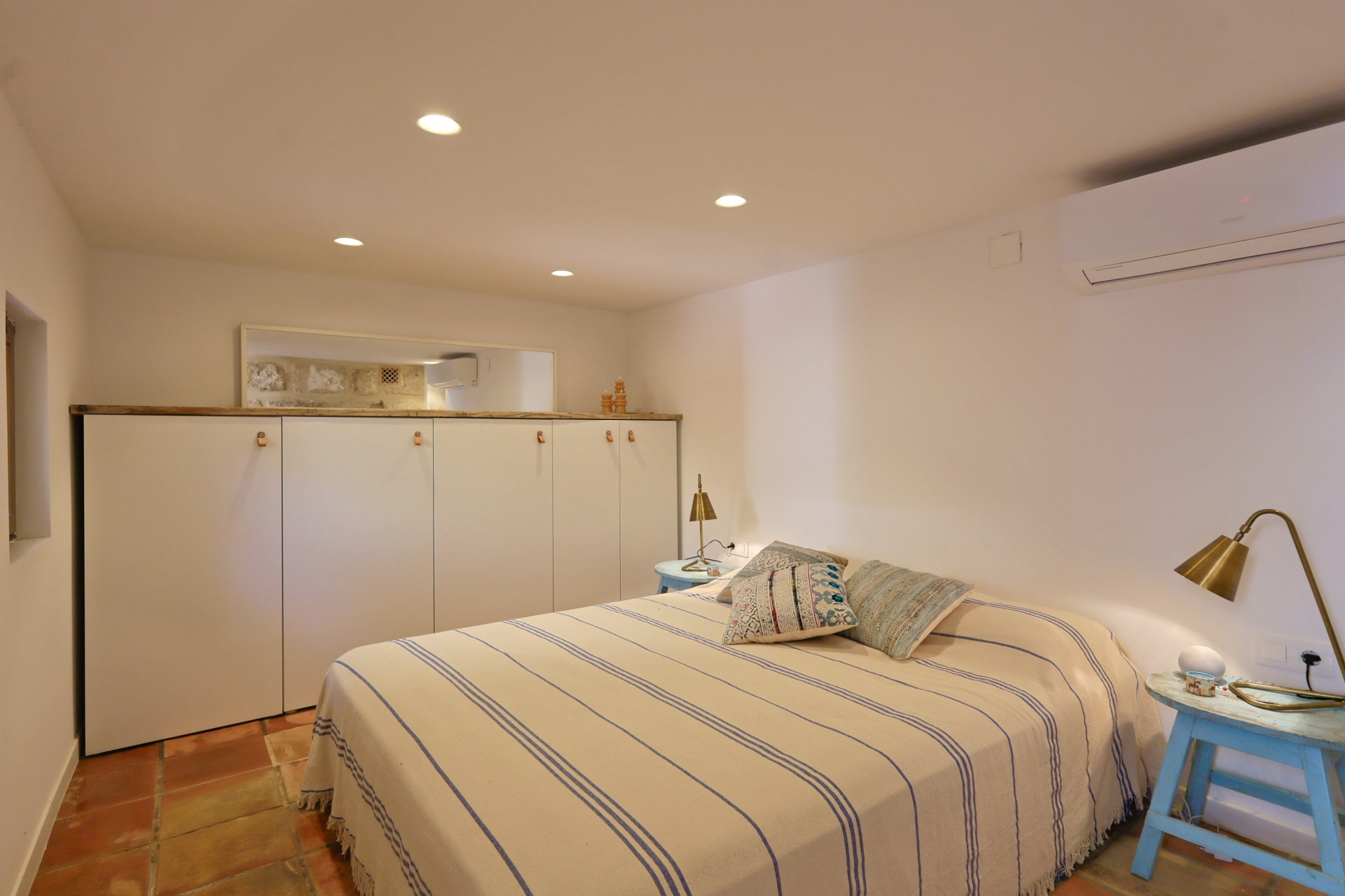 Dormitorio luminoso con suelo de terracota, cama con cubierta a rayas y armario empotrado blanco con detalles en madera.