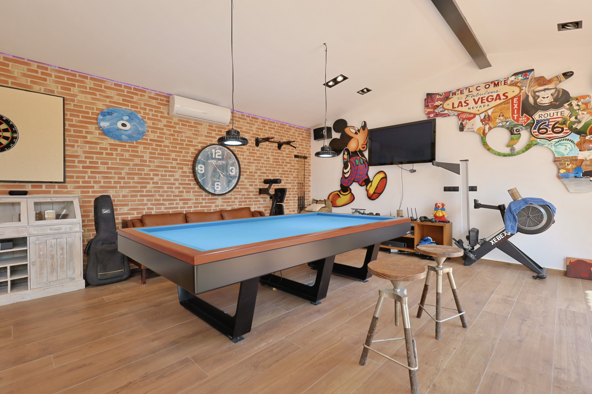 Sala de juegos con mesa de billar azul, pared de ladrillo visto y decoración temática.