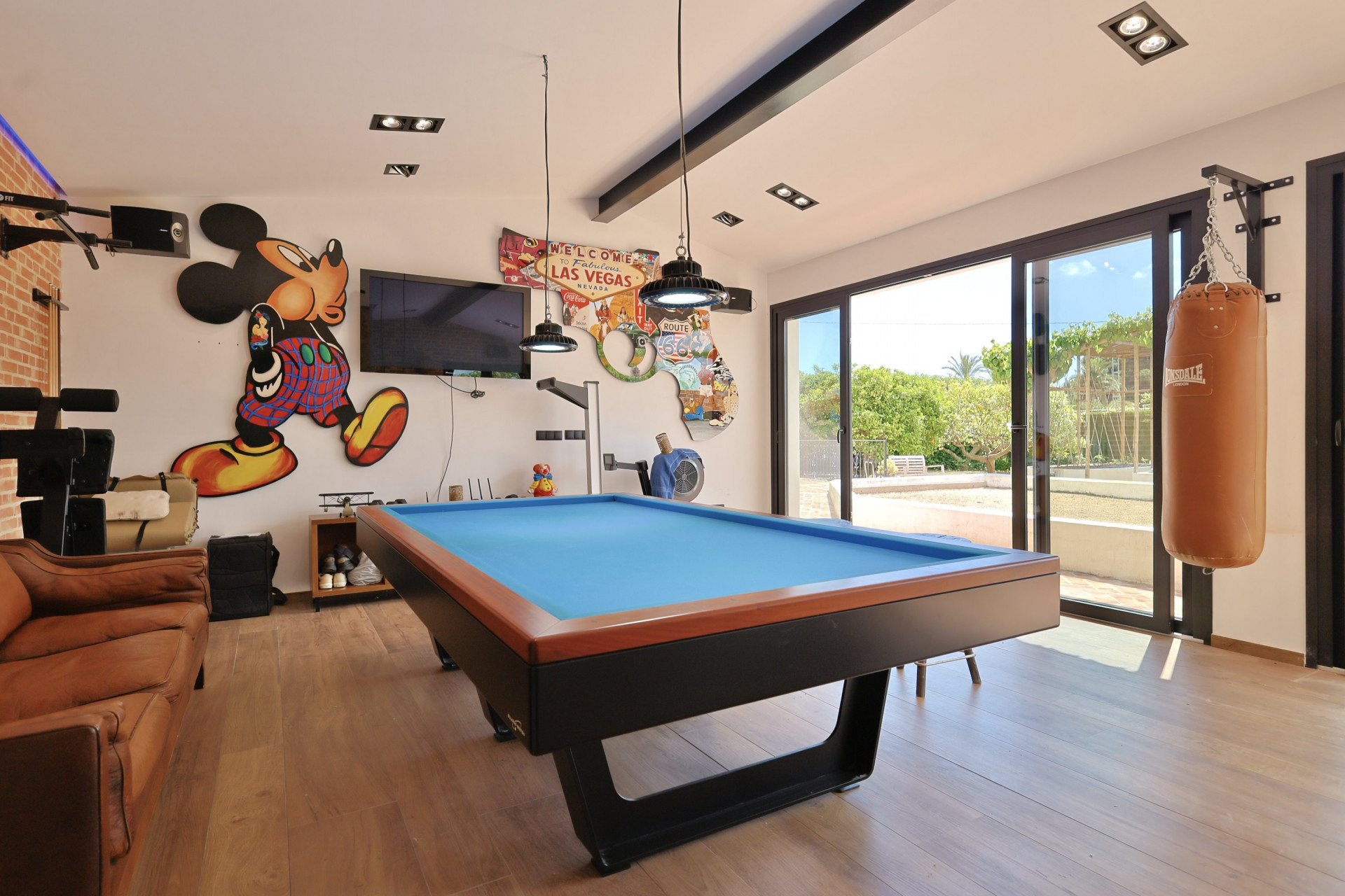 Sala de juegos con mesa de billar azul y decoración temática, incluye un saco de boxeo y acceso al jardín.
