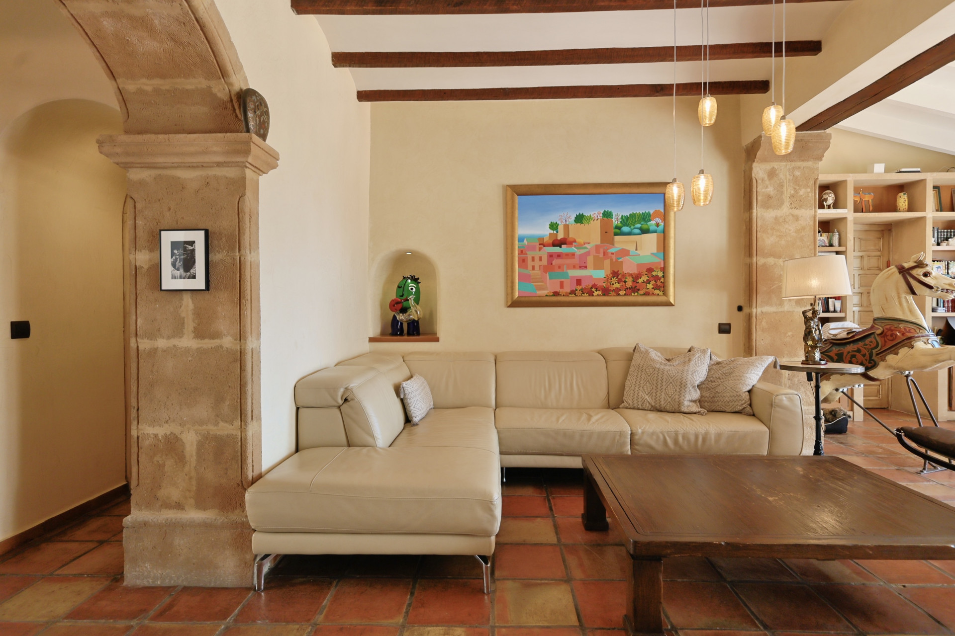 Salón con sofá de cuero, mesa de centro rústica y suelo de terracota. Detalle de arco de piedra.