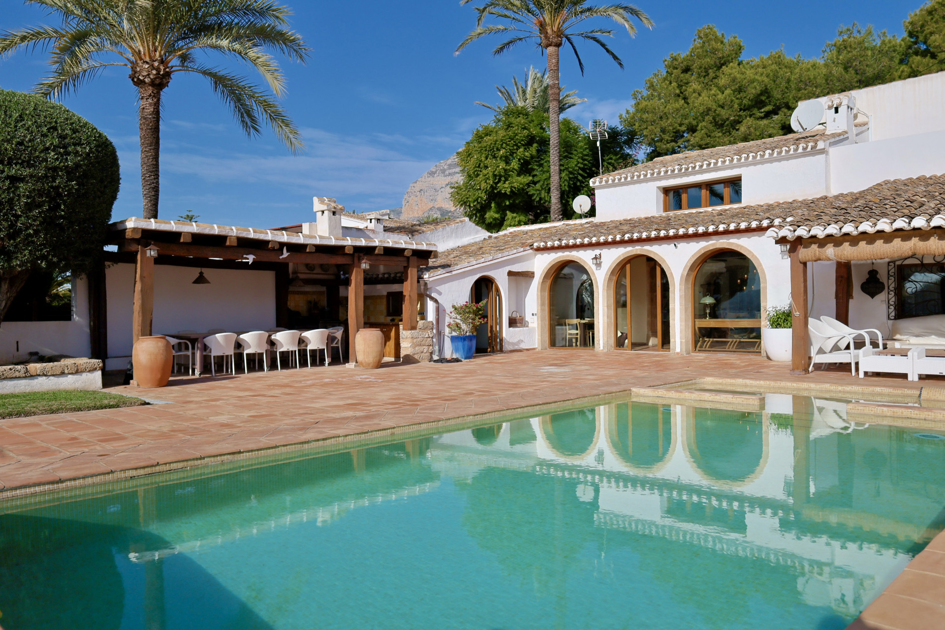 Vista de la piscina turquesa reflejando la arquitectura de arcos y la pérgola con comedor exterior.