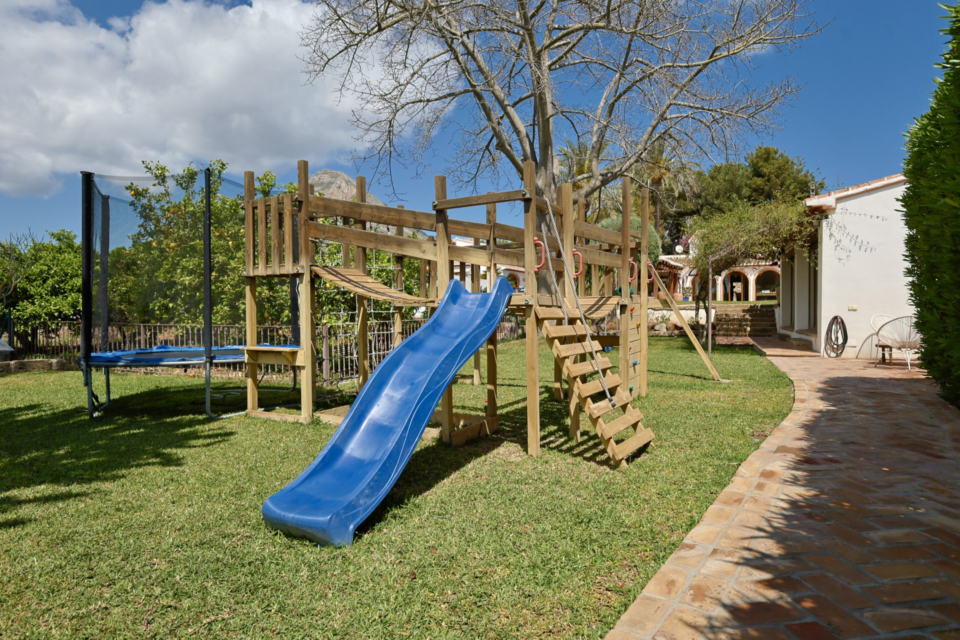 Zona de juegos con estructura de madera, tobogán azul y trampolín en el jardín.