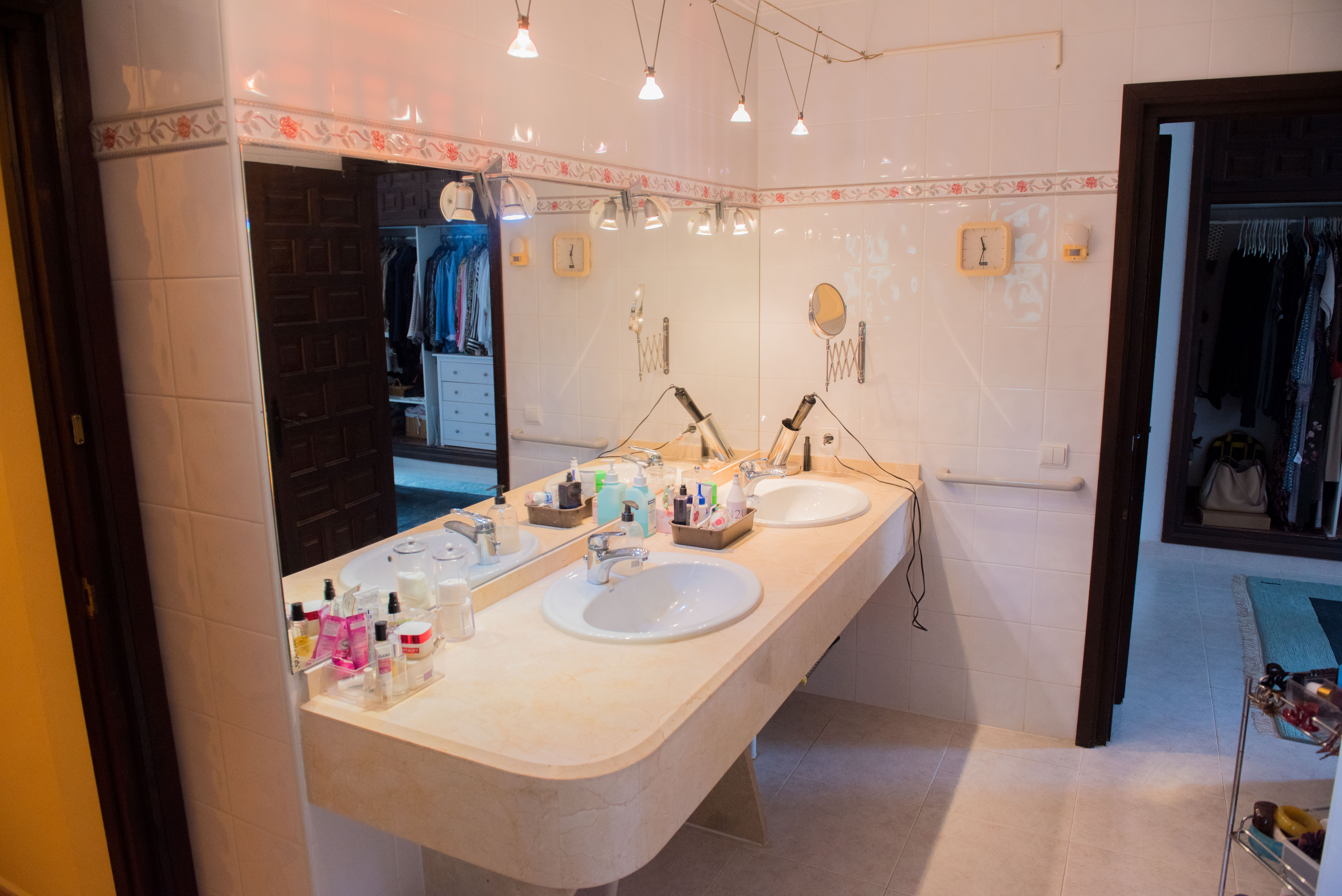 Baño con encimera de mármol crema, lavabo de doble seno y gran espejo que refleja un vestidor organizado con carpintería de madera oscura.