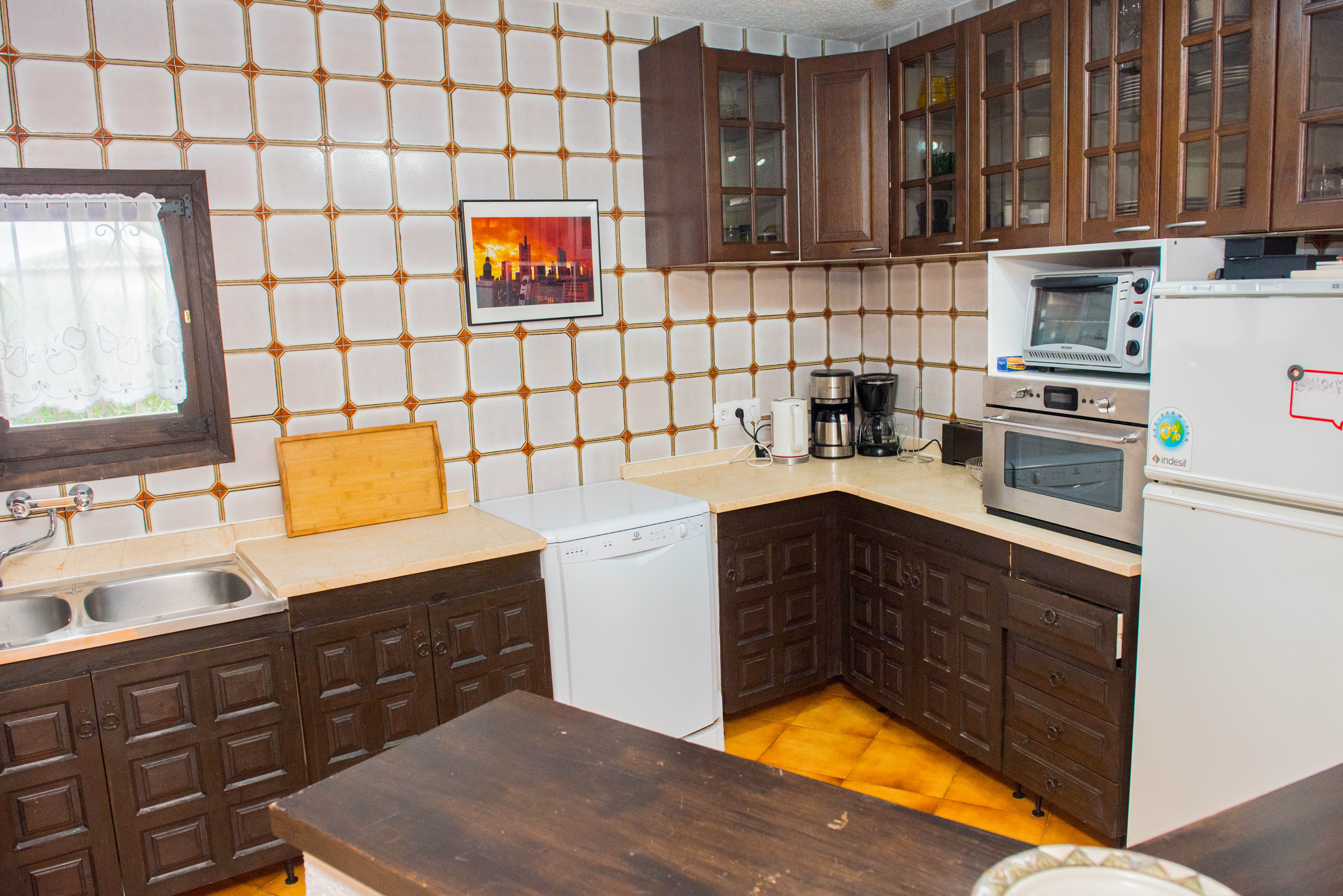 Cocina rústica con armarios de madera oscura tallada, encimera de tono crema y azulejos con motivos geométricos. Incluye lavavajillas blanco y horno integrado.