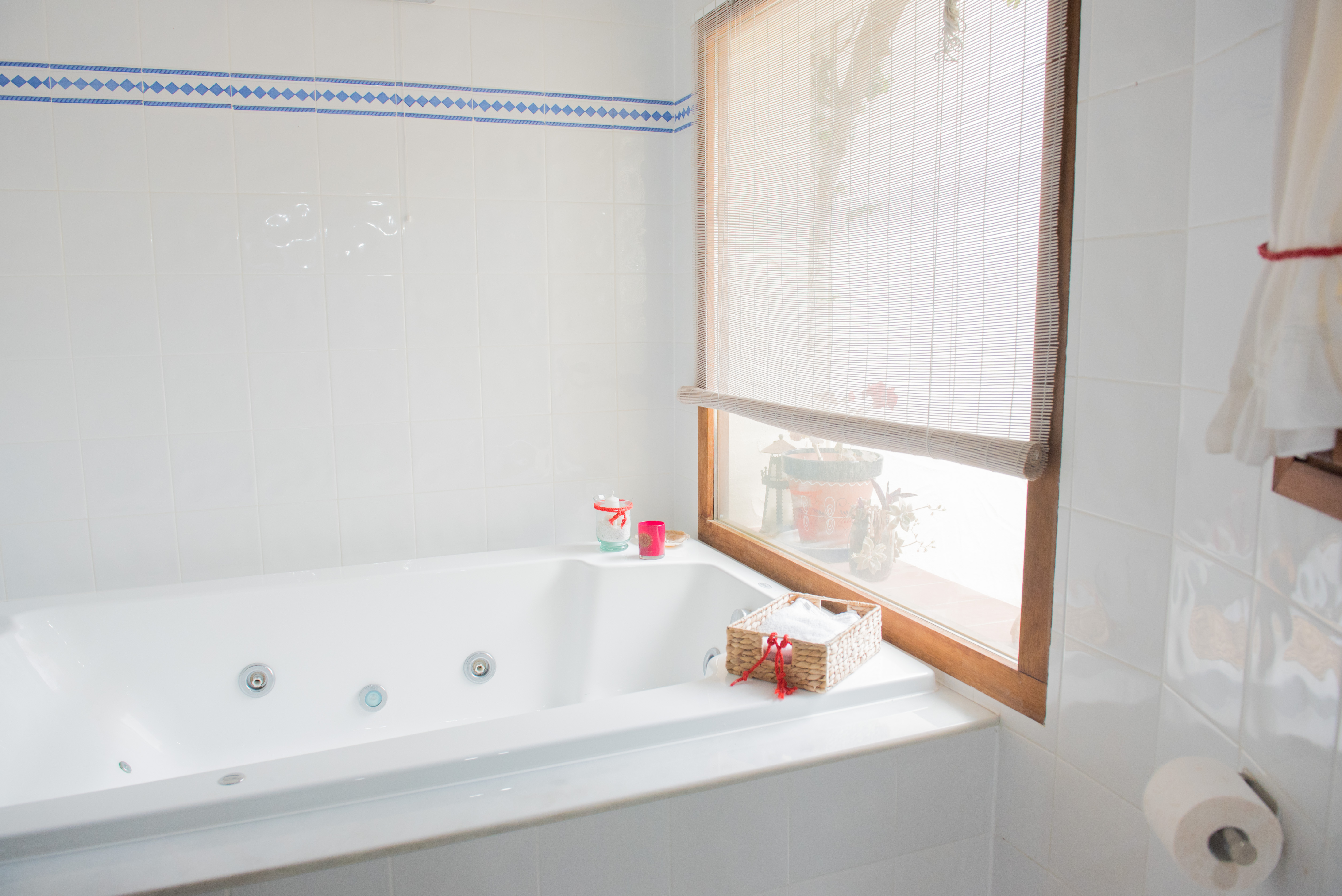 Cuarto de baño con bañera de hidromasaje integrada, paredes alicatadas en blanco con detalle de cenefa azul y ventana de madera con persiana de bambú.