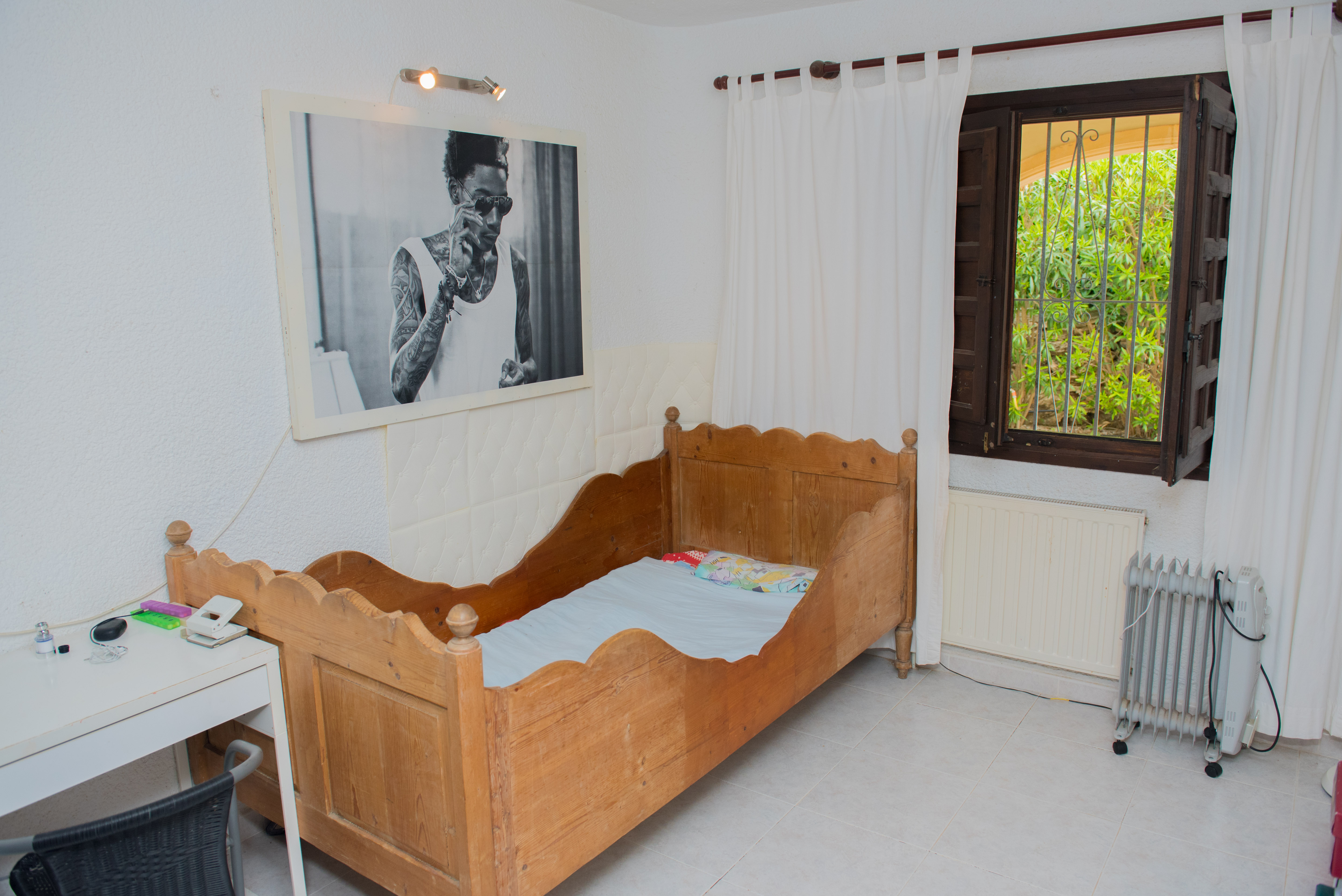 Dormitorio con cama de madera rústica de diseño ondulado, escritorio blanco y ventana de madera oscura con rejas. Suelo de baldosas claras y luz natural.