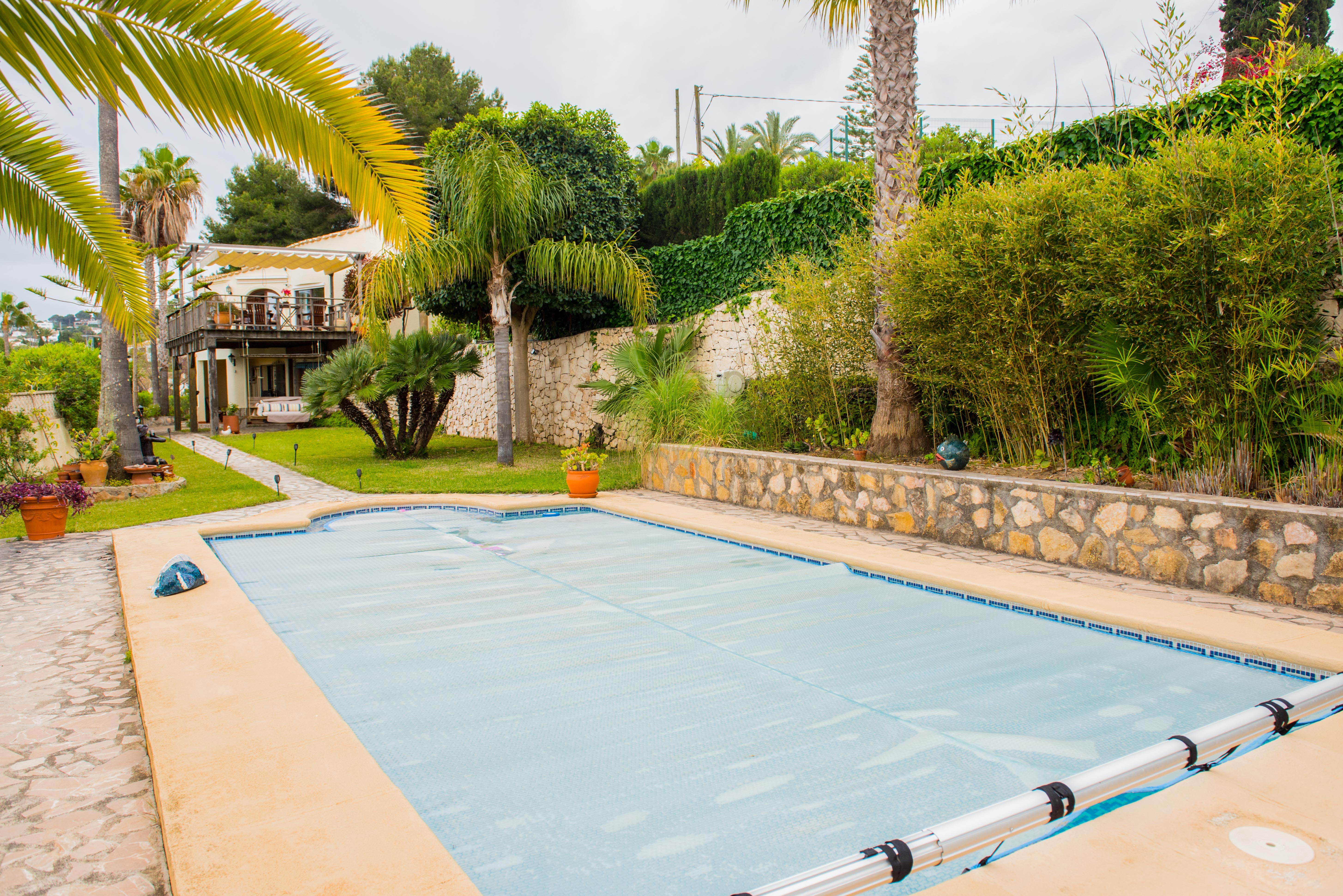 Piscina exterior con cubierta térmica azul y rodillo metálico, rodeada de muros de piedra natural, palmeras y vegetación mediterránea consolidada frente a la vivienda.