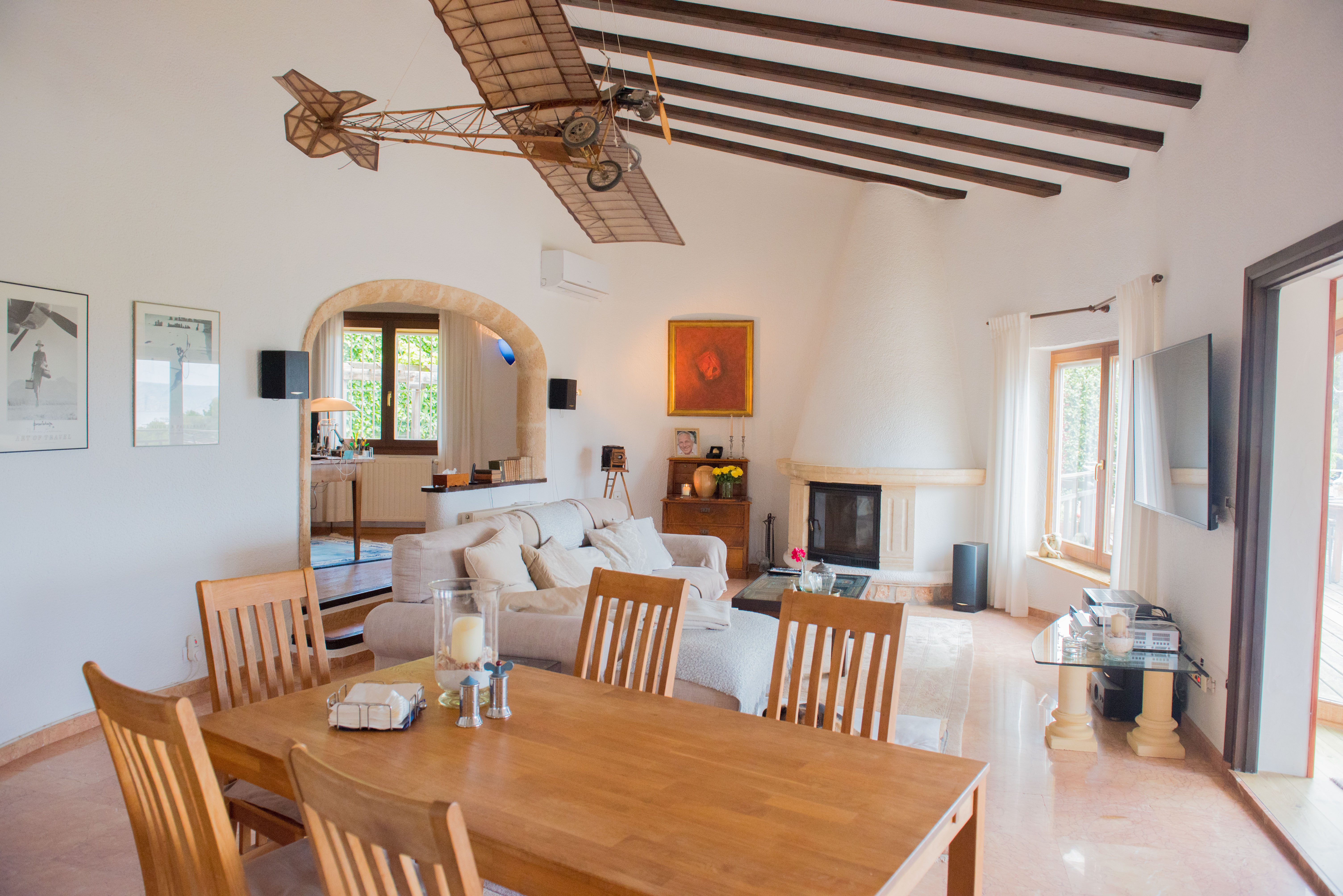 Salón comedor de techos altos con vigas de madera, chimenea cónica y arco de piedra que comunica con el despacho. Destaca la luz natural y el suelo de mármol pulido.