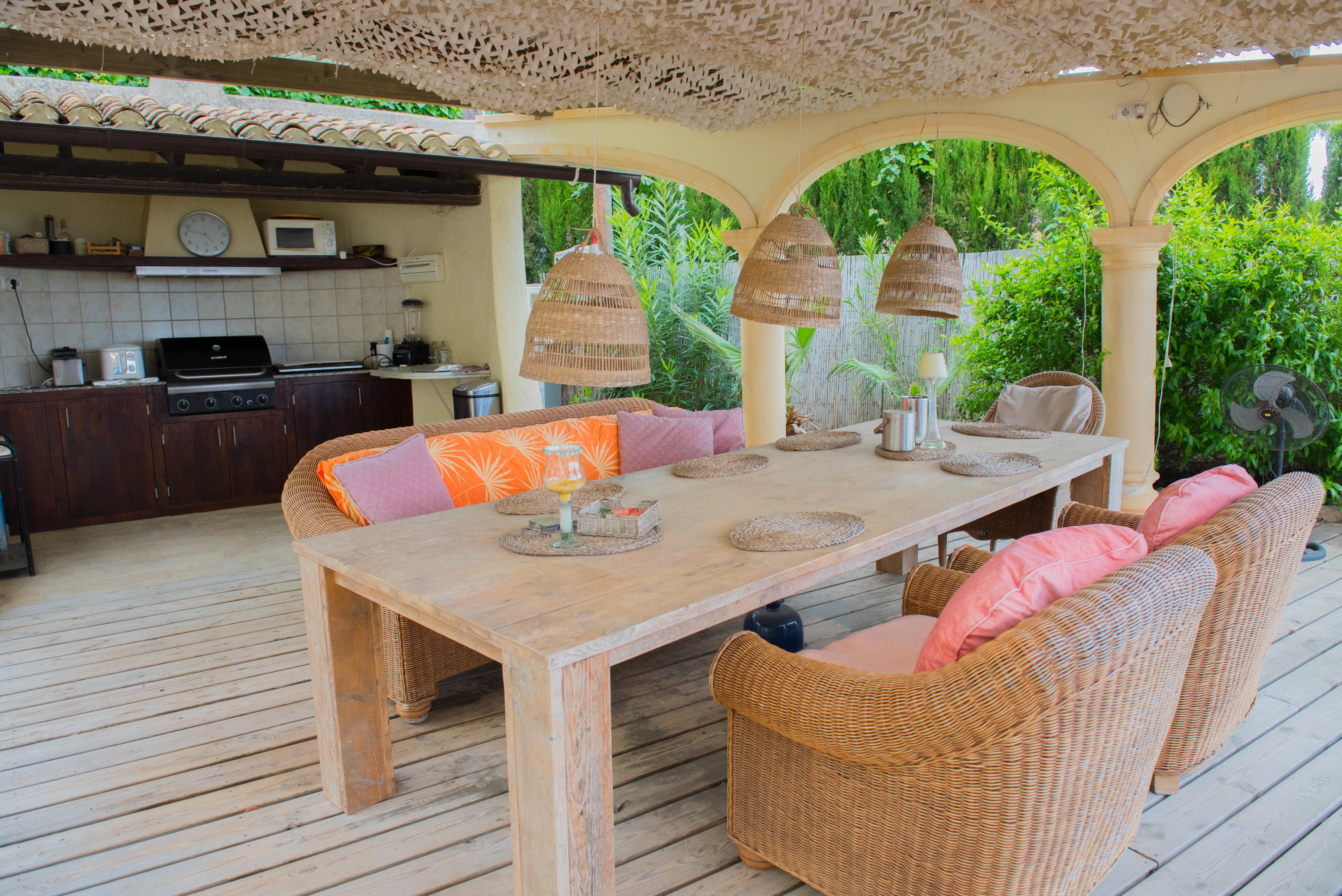 Terraza cubierta con cocina de verano equipada, mesa de madera maciza, sillones de mimbre y techado decorativo sobre suelo de tarima de madera.