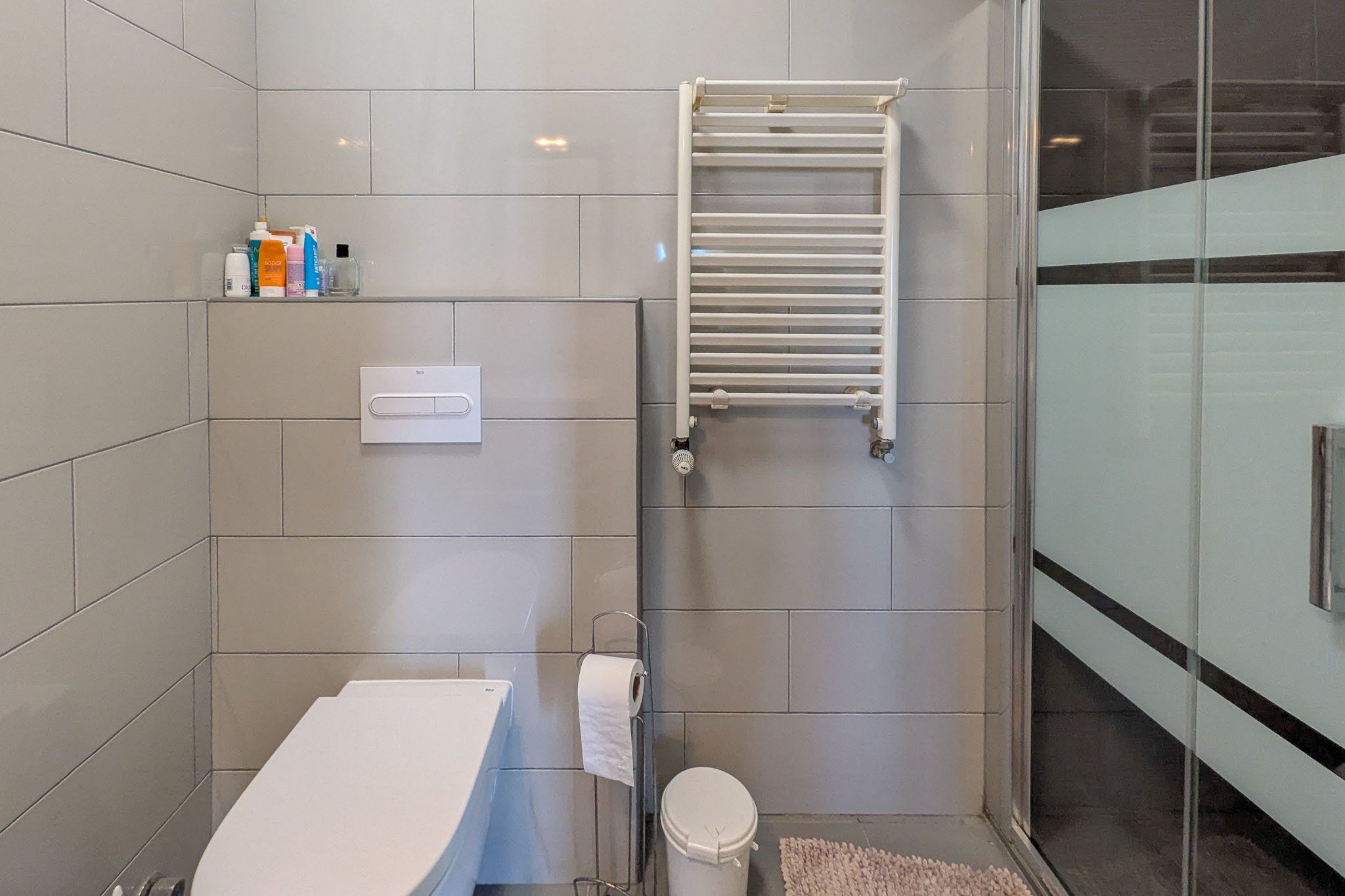 Baño moderno con inodoro suspendido, revestimiento de gres gris, radiador toallero blanco y mampara de ducha de cristal con franja de privacidad traslúcida.