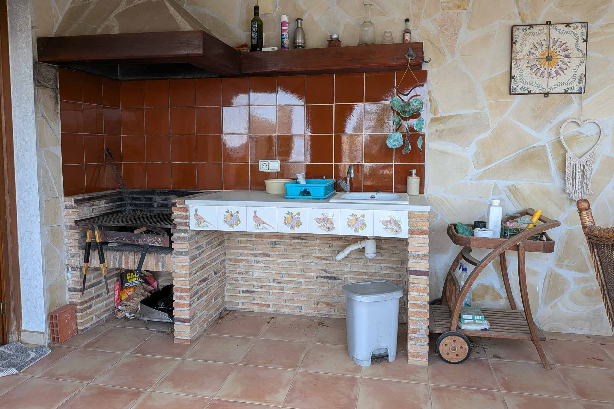 Cocina de verano exterior con barbacoa de obra, fregadero integrado y encimera decorativa sobre muros de ladrillo visto y paredes de piedra natural.