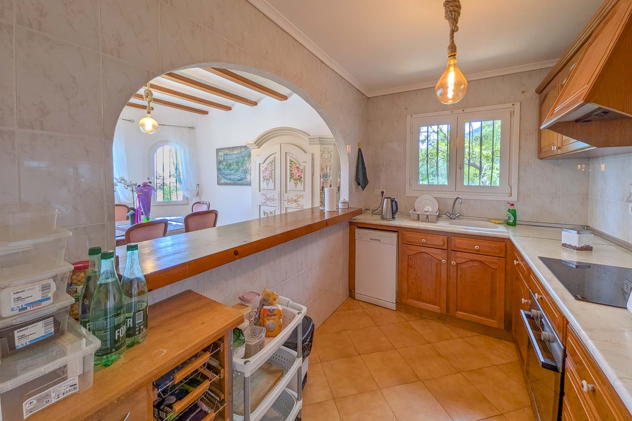 Cocina semi-abierta con mobiliario de madera, encimera de madera maciza y arco comunicante al comedor con vigas a la vista y suelos cerámicos.