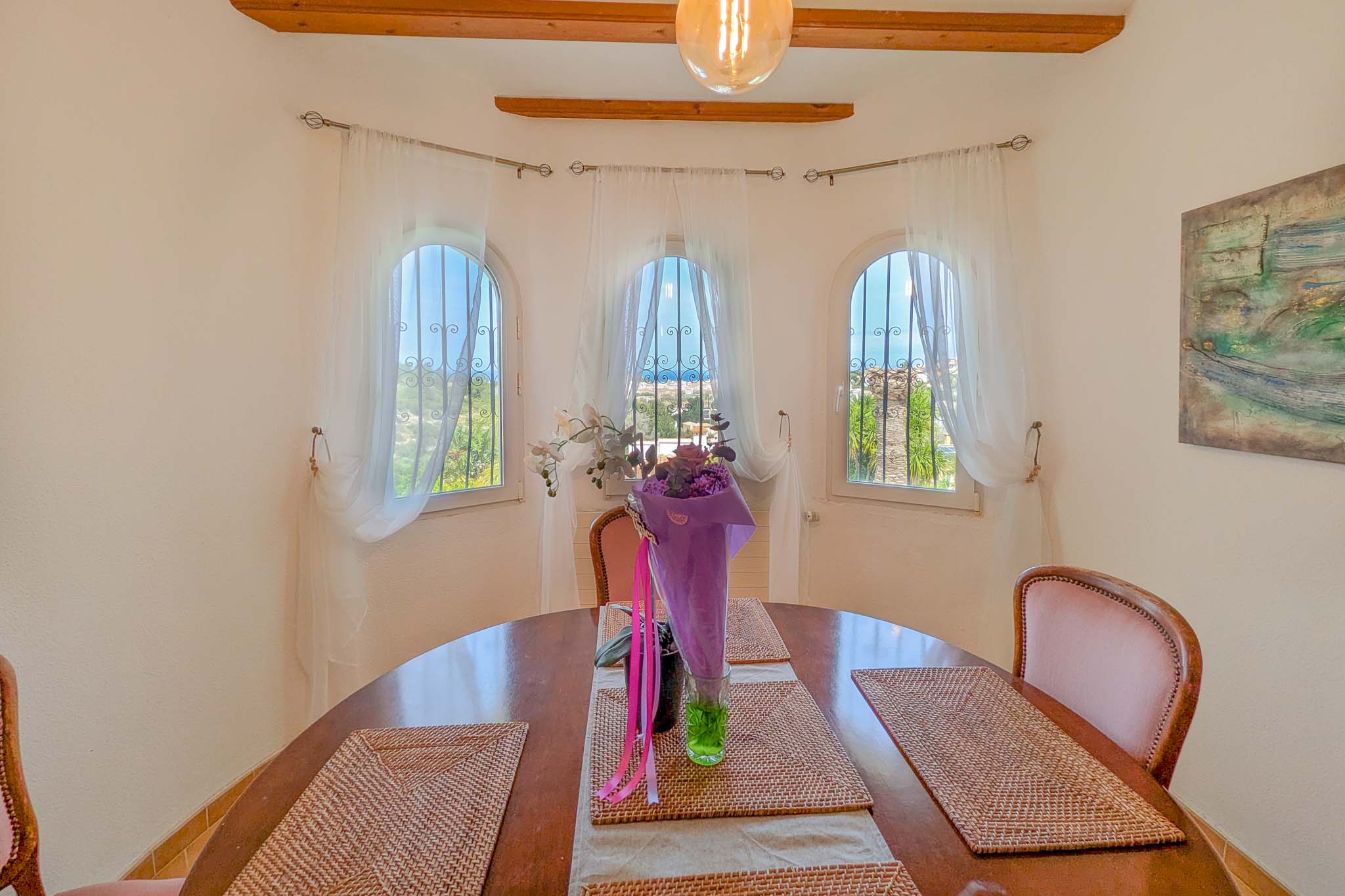 Comedor con mesa circular de madera y tres ventanales de arco con vistas al Mediterráneo. El espacio cuenta con vigas de madera vista y abundante luz natural.