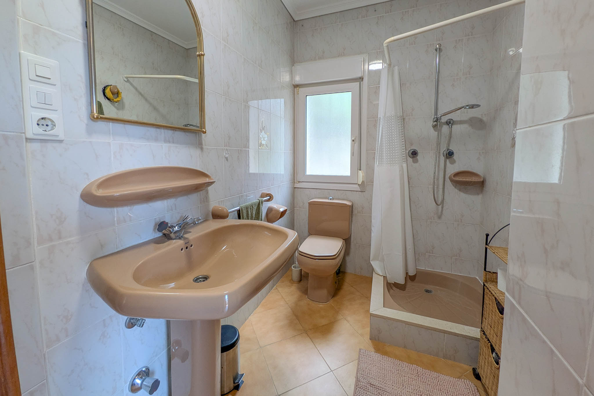 Cuarto de baño con sanitarios en tono beige, plato de ducha con cortina, paredes alicatadas y ventana exterior que aporta luz natural.