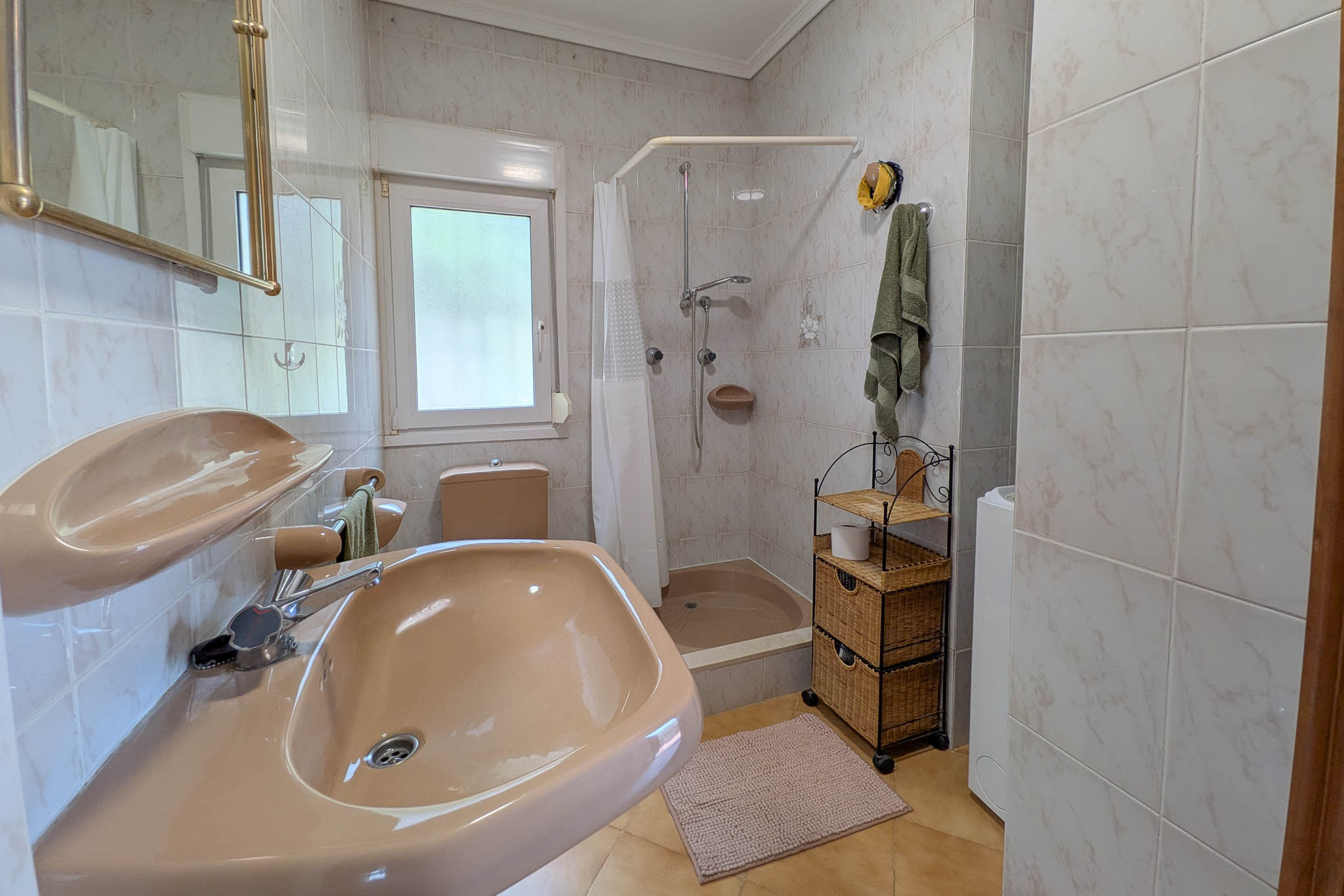 Cuarto de baño funcional con sanitarios en tono salmón, plato de ducha con cortina, paredes alicatadas y ventana de climalit para ventilación natural.