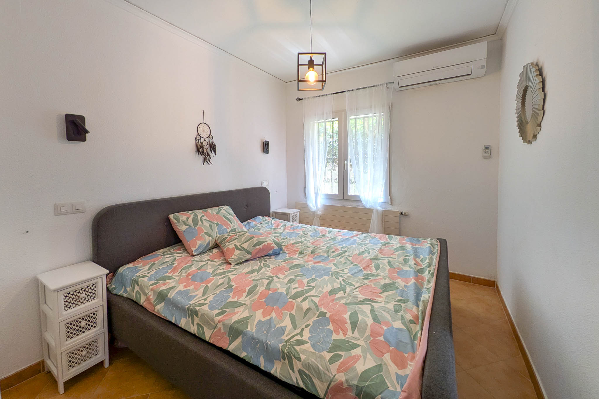 Dormitorio con cama de matrimonio, cabecero tapizado en gris, aire acondicionado split y suelos de gres. La estancia cuenta con iluminación natural y una lámpara colgante de estilo industrial.