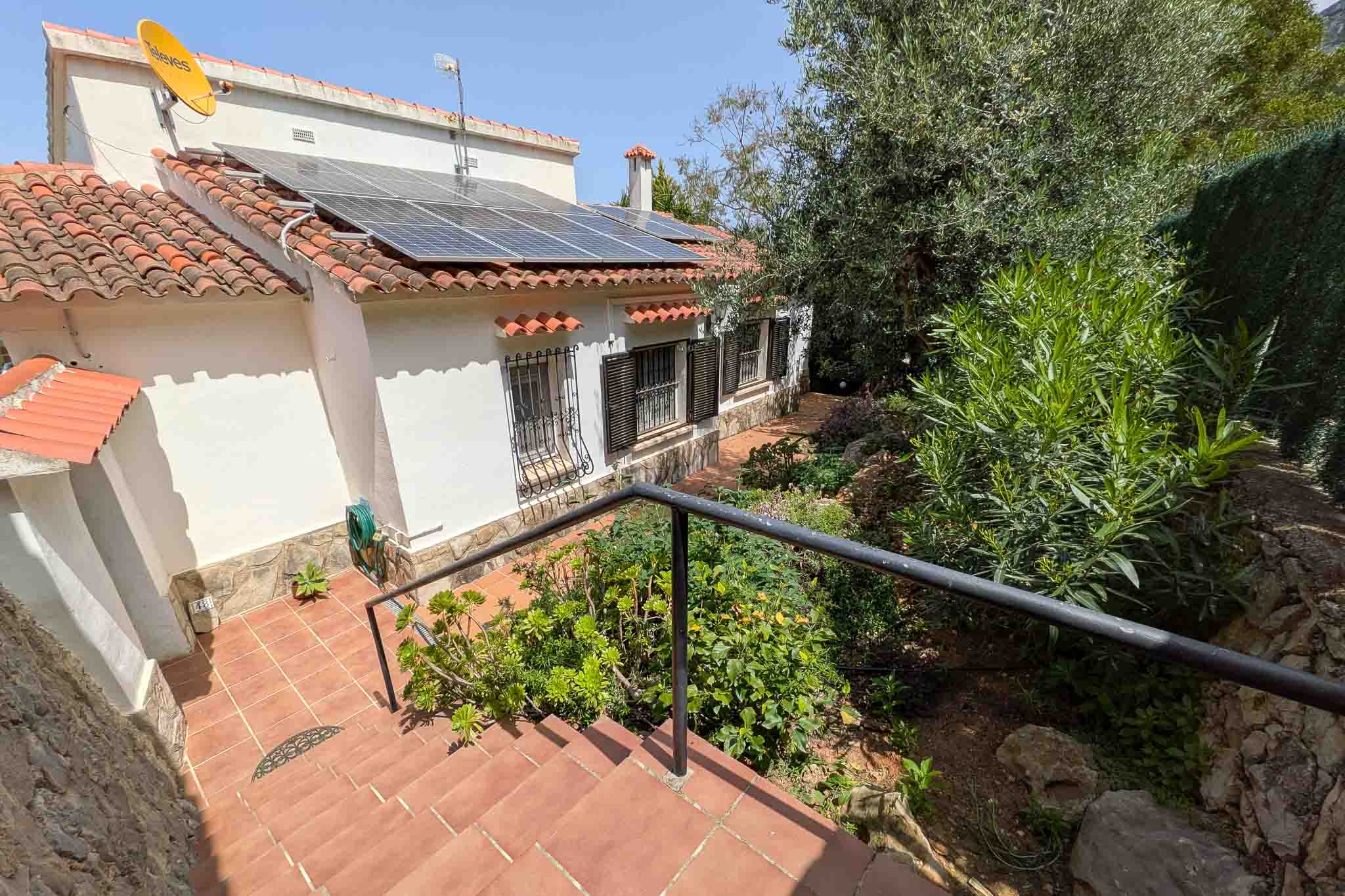 Exterior de la vivienda con fachada blanca, paneles fotovoltaicos en el tejado y acceso mediante escalera de terracota rodeada de vegetación mediterránea.